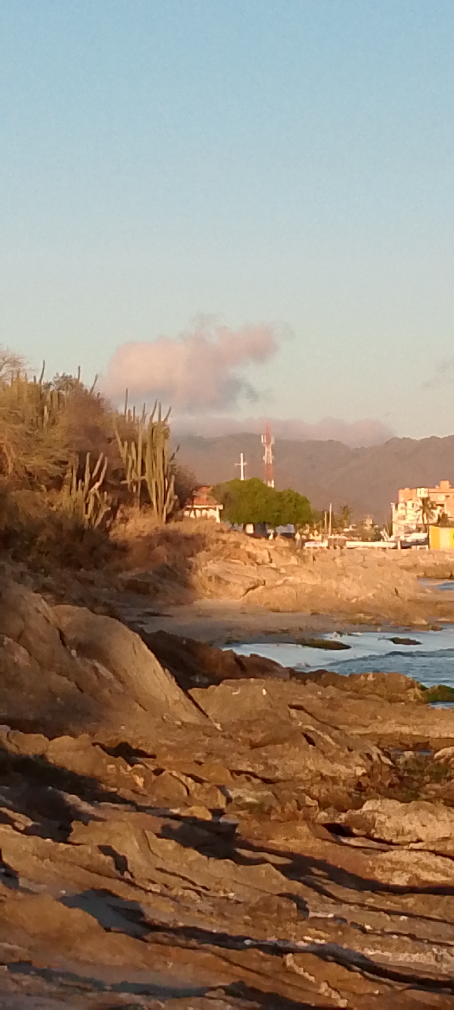The image depicts a coastal landscape in Juan Griego, Venezuela, showcasing a rocky shoreline in the foreground. The middle ground features dry vegetation including cacti, a white Christian cross, and a communication tower. In the background, there are hazy mountains and buildings indicating urban development. The sky is light blue with some light clouds. The photo was likely taken during the late afternoon or early morning given the warm, golden light.