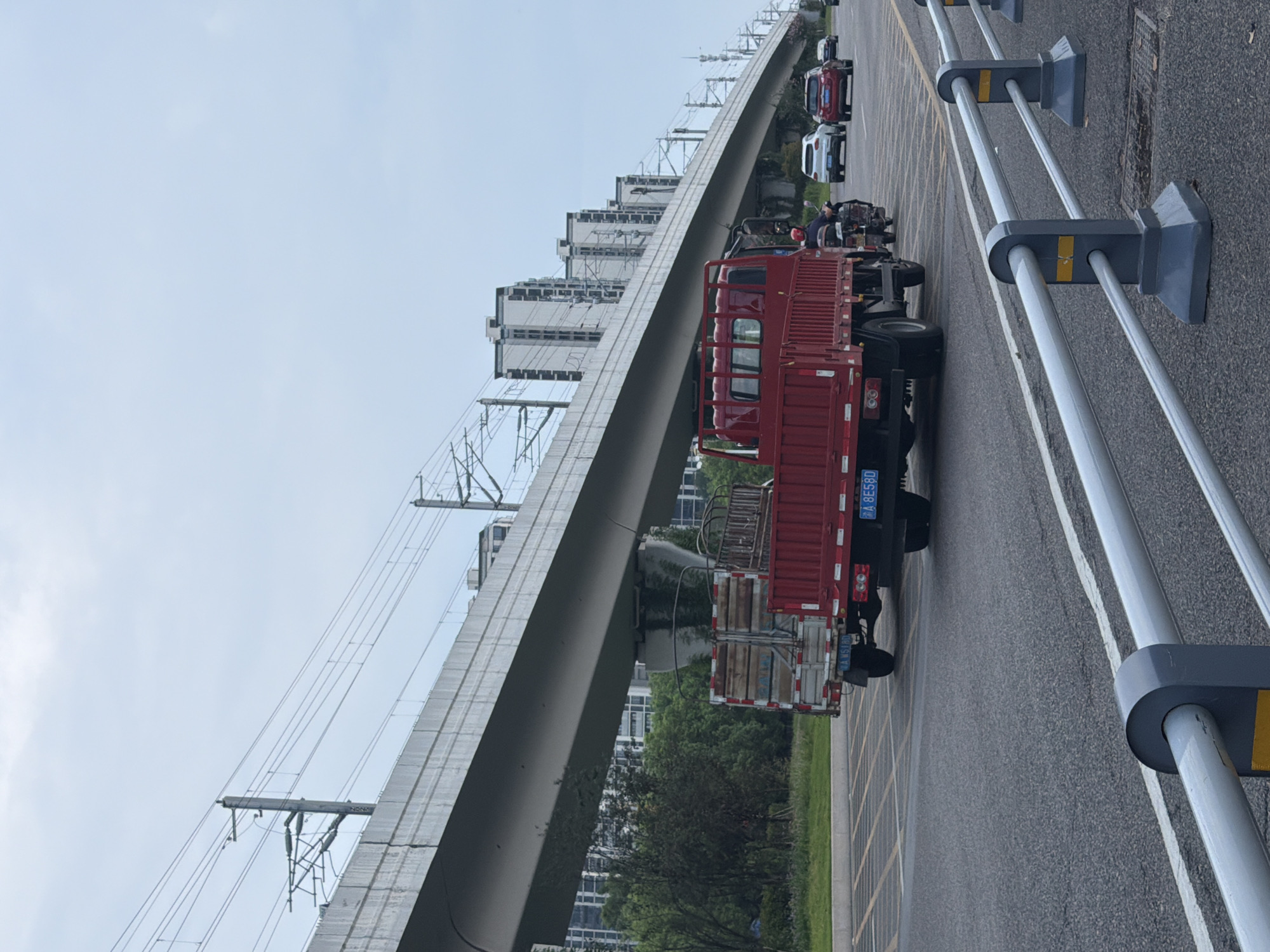The image depicts a street scene in Jiangjin, China, with an elevated railway line and buildings in the background. In the foreground, a red flatbed truck is being driven, towing a trailer. The truck has a license plate that reads "A 8E58D". A motorcycle with a rider is visible next to the truck. Further down the road, there are several cars, including a white SUV and a red car. The road is paved, and there are guardrails along the side. The sky is overcast, and there are overhead power lines associated with the railway. Trees and greenery are present between the road and the buildings.