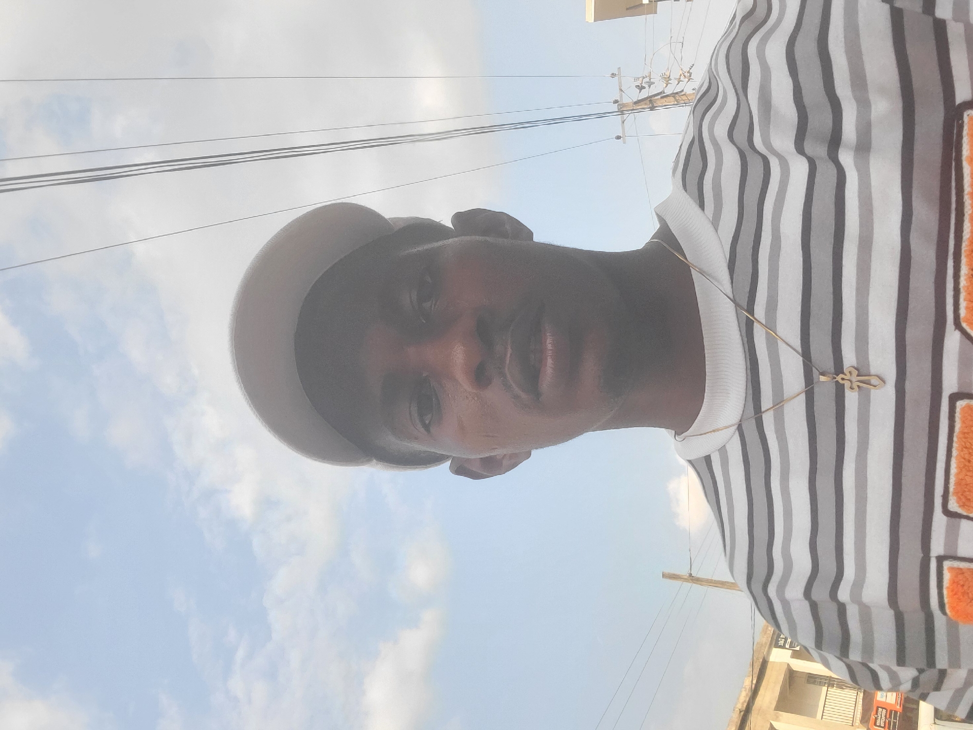 The image shows a man posing for a selfie, with the camera held at a low angle, pointing upwards towards his face. He is wearing a striped t-shirt with white, gray, and black lines, and a light-colored cap. A gold chain with a cross pendant is visible around his neck. The background consists of a bright, overcast sky with some clouds and power lines. The orientation of the image is rotated.
The man's face is partially obscured by the angle and lighting. He appears to be outdoors, possibly in an urban or suburban environment, given the presence of power lines and a glimpse of a building in the lower right corner. The time of day is likely daytime, as indicated by the bright sky. The overall mood is casual and confident. No specific activity or event is depicted, other than the act of taking a selfie.
The image does not contain any visible text that is legible. The location context provided is Egbagu, Nigeria.