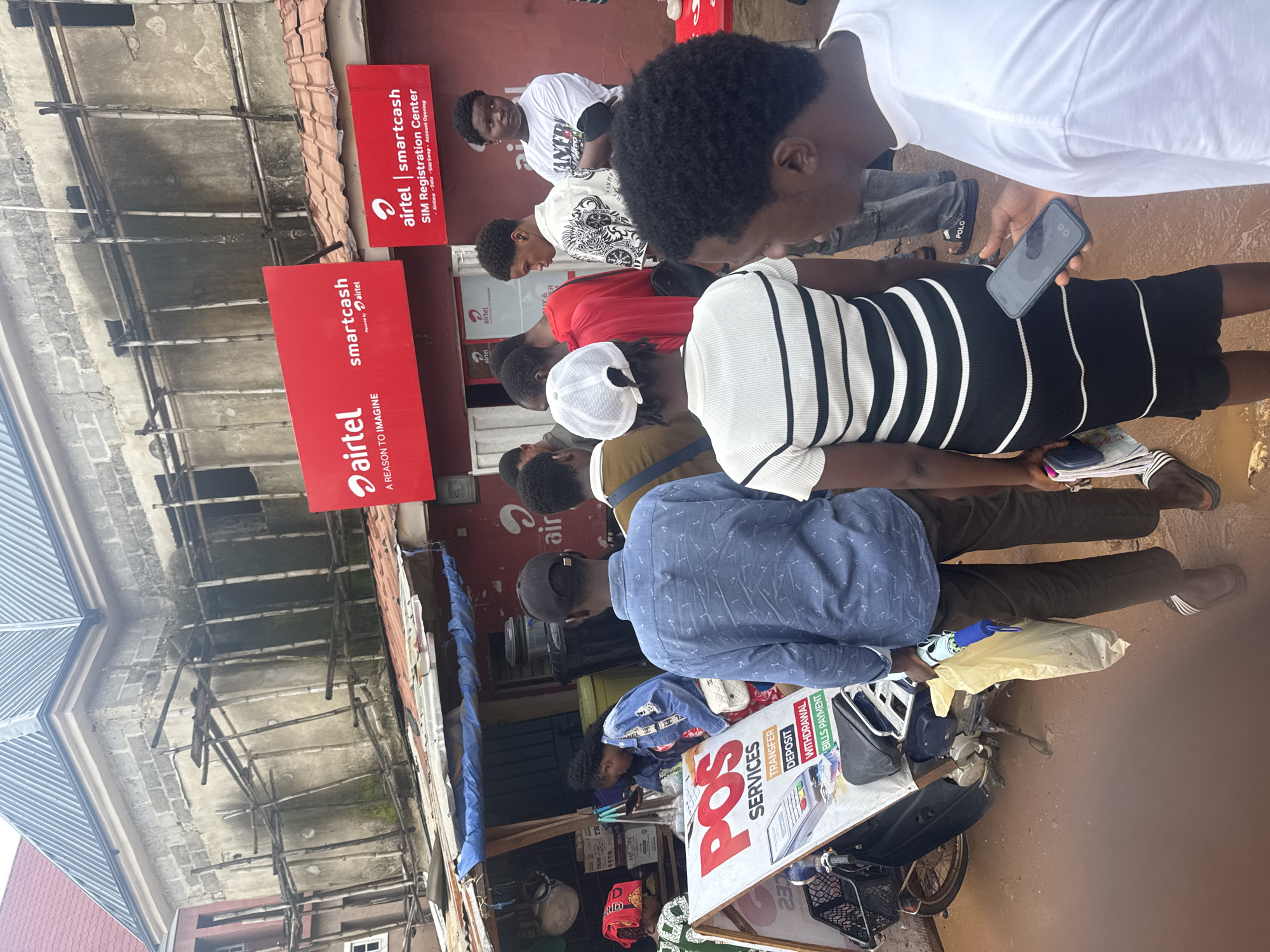 In Ihiagwa, Nigeria, a group of people are gathered outside a business establishment with red signage displaying "Airtel" and "Smartcash." The building behind them appears to be under construction, with scaffolding visible on its exterior. In the foreground, a POS services sign is displayed on an A-frame, listing services such as transfer, deposit, withdrawal, and bills payment. Several individuals, including men and women of various ages, are seen interacting or standing in close proximity. One person is holding a smartphone, and another is carrying a yellow bag and a blue umbrella. The ground appears wet, suggesting recent rain. The overall scene depicts a busy urban environment with commercial activity and ongoing development.