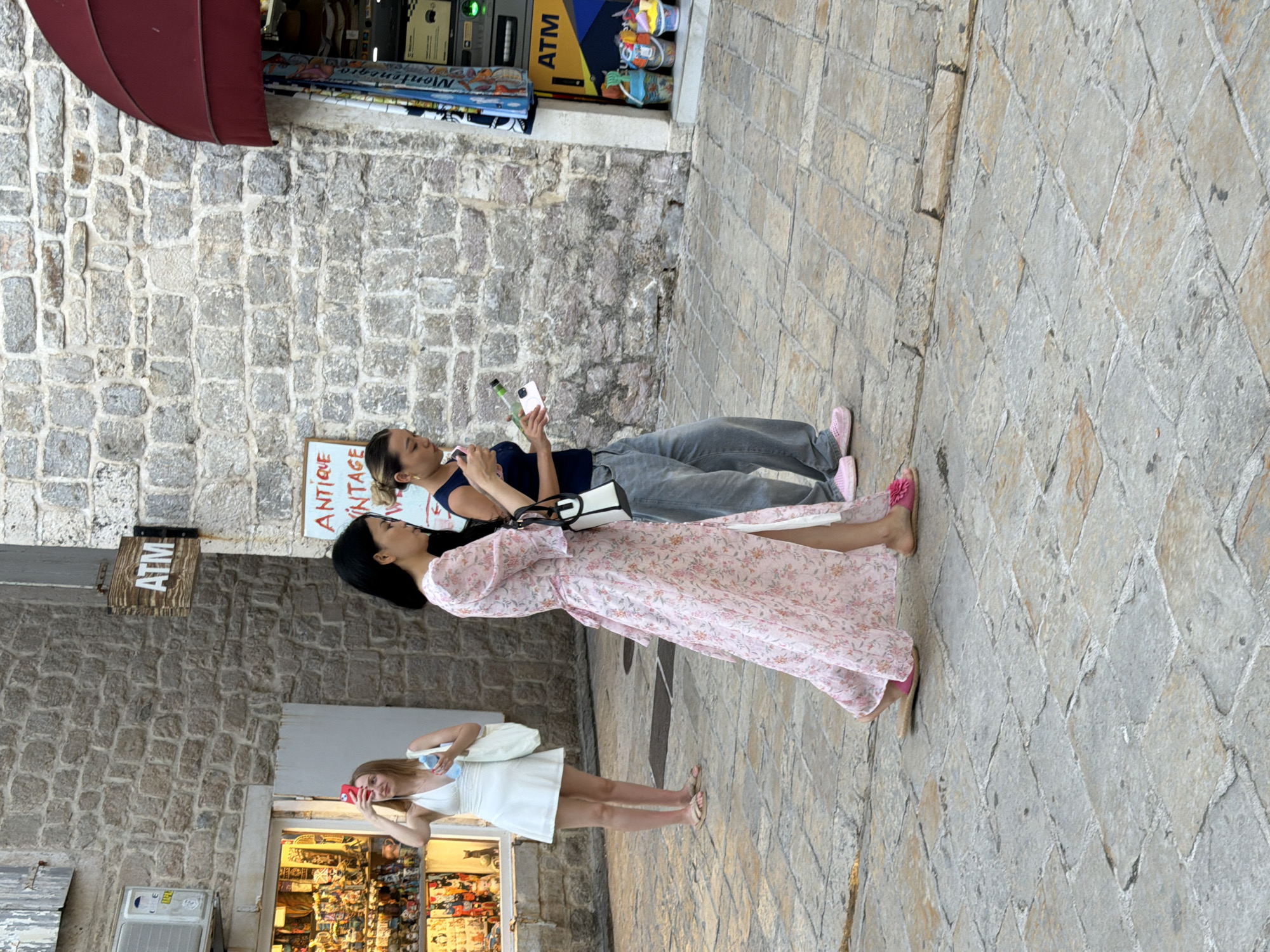 In Budva, Montenegro, a woman in a white dress stands in front of a storefront, holding a phone to her face as if taking a picture. To her right, two women walk down a cobblestone street. The woman in the foreground wears a long, pink floral dress and carries a white purse. The woman behind her, in a dark blue top and grey jeans, holds a phone and a green bottle. Behind them, an ATM is visible next to a shop with red awnings. The buildings are made of stone, and the street is paved with square stones.