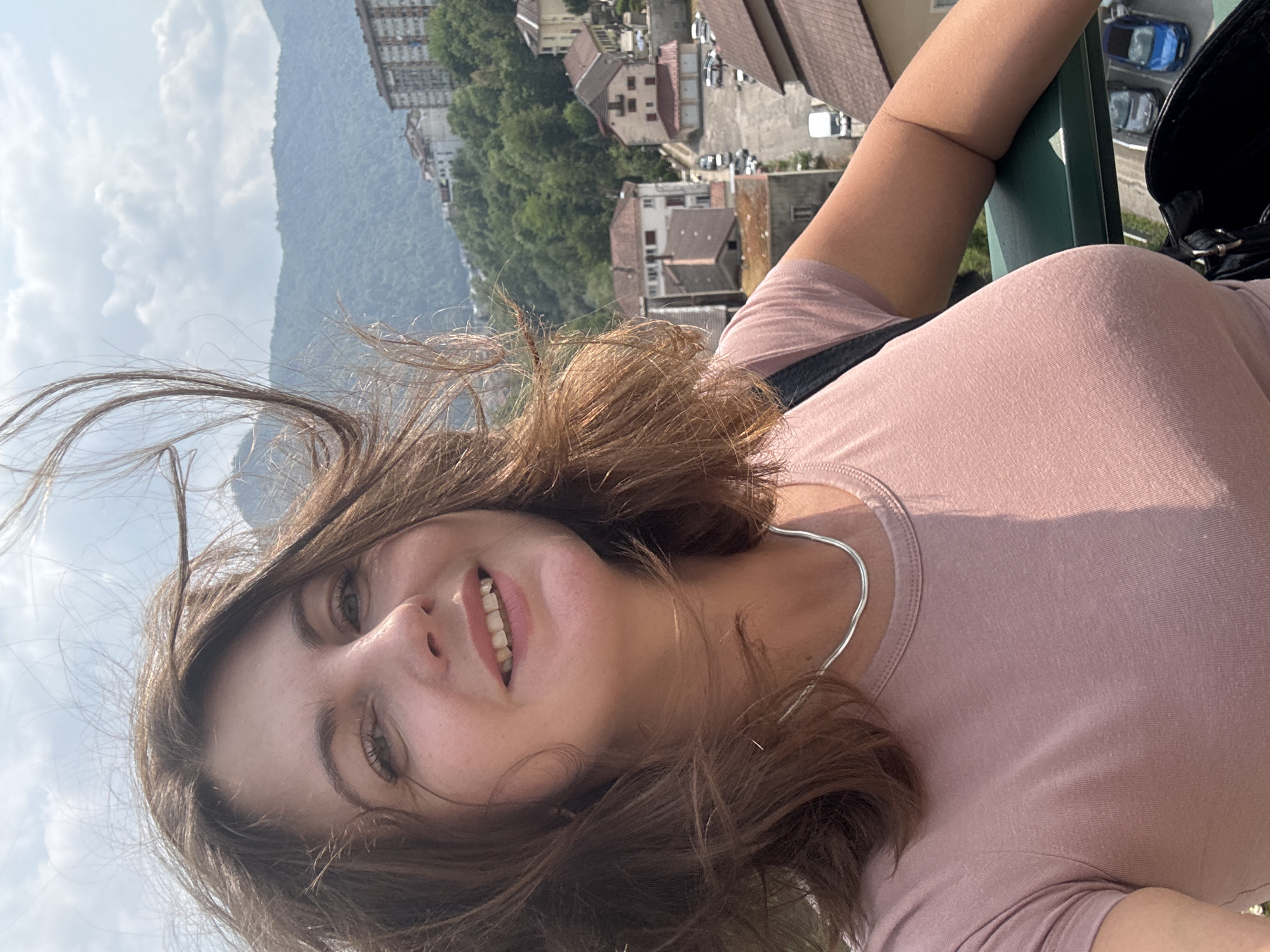A woman is taking a selfie outdoors, with a city and mountainous landscape in the background. She has medium-length brown hair that is blowing in the wind, and she is wearing a pink t-shirt and a silver necklace. The city appears to be Saint-Claude, France, characterized by its red-tiled roofs and buildings nestled on a hillside. The sky is partly cloudy.