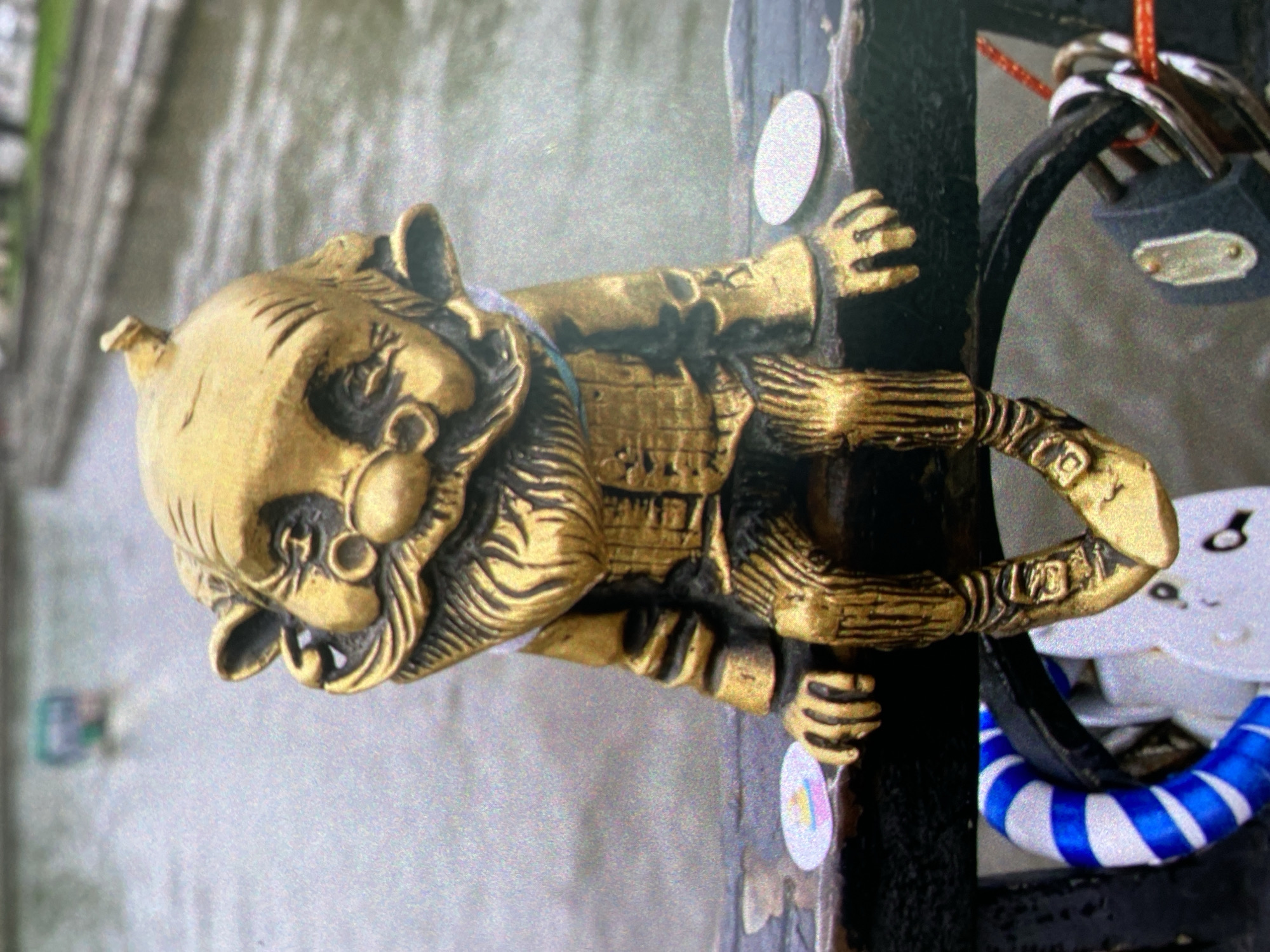 A metal sculpture of a mythical creature, possibly a house gnome or similar folklore being, is affixed to a dark-colored post or vertical structure. The sculpture is primarily golden in color with dark lines and detailing, giving it a somewhat antique appearance. The creature has a prominent nose, tusks, and ears, wearing a suit-like garment. The sculpture's hands appear to grasp or hold the post.
To the right of the sculpture, there is a black object with padlocks attached, suggesting it could be a bridge railing where love locks are placed. A blue and white striped ribbon or decoration is also visible.
The blurred background suggests the sculpture is located in a public place, possibly near a body of water or a pedestrian walkway. Given the style of love locks and the folklore figure, it's plausible this is located in a touristy area, though this is speculative.