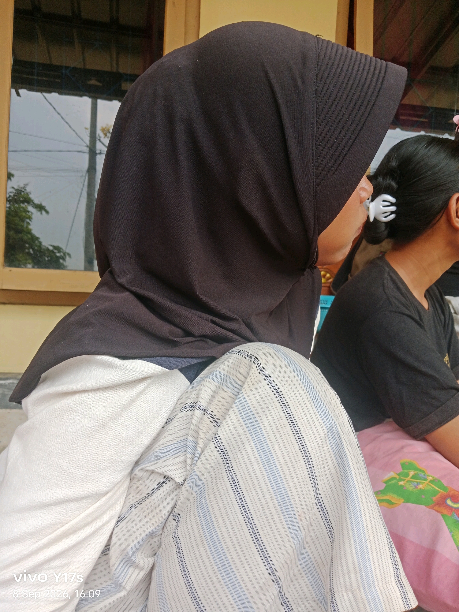 A medium shot shows two individuals sitting. The person in the foreground, facing right, wears a dark brown hijab and a cream-colored shirt with a striped pattern. Their right hand is partially visible. To the right, another person, facing away, has dark hair held by a white clip and wears a dark gray t-shirt. They are seated next to a pink blanket with a green giraffe design. The background features a window with a wooden frame, reflecting an outdoor scene with trees and power lines. The location is identified as Rama Dewa, Indonesia, with a timestamp of 8 Sep 2020, 16:09.