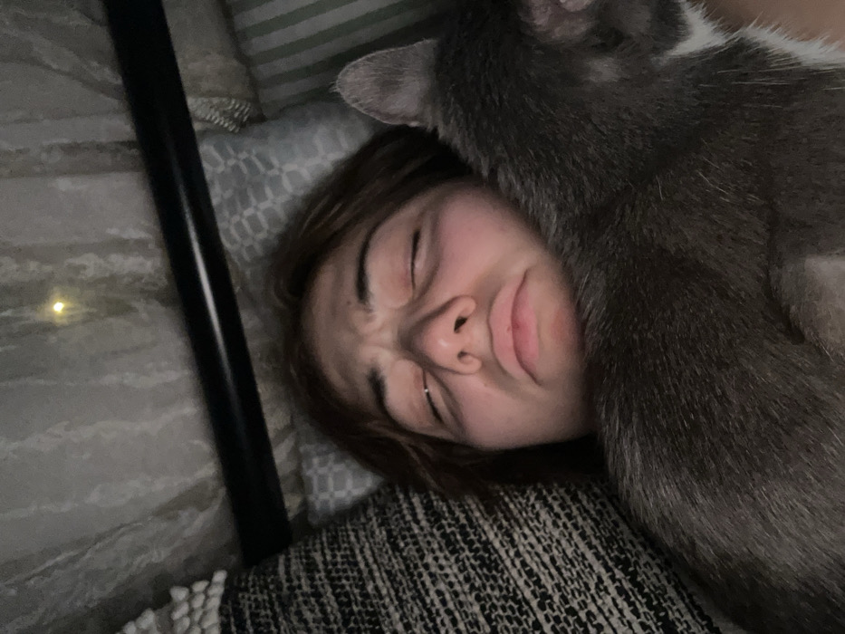 A young woman lies on patterned pillows, her eyes squinted and lips pursed in an exaggerated expression. A large, gray cat is sprawled on her, its fur covering part of her face. A black pole is visible on the left. The scene is shot at a close, high angle, with muted lighting, creating a cozy yet humorous mood.