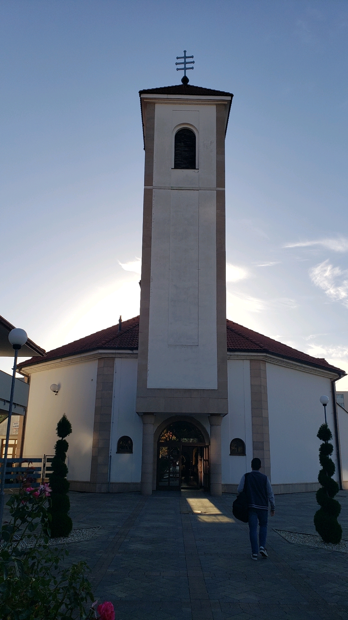 The image shows the exterior of a church in Prešov, Slovakia. The church has a tall, white bell tower with a cross on top and a large arched entrance. The main building is white with a red tiled roof and stone accents. In front of the church, a person is walking away from the camera towards the entrance, carrying a dark bag. The ground in front of the church is paved with cobblestones. There are decorative bushes on either side of the path and some flowers in the foreground. The sky is blue with some wispy clouds, and the sun is setting, casting long shadows.