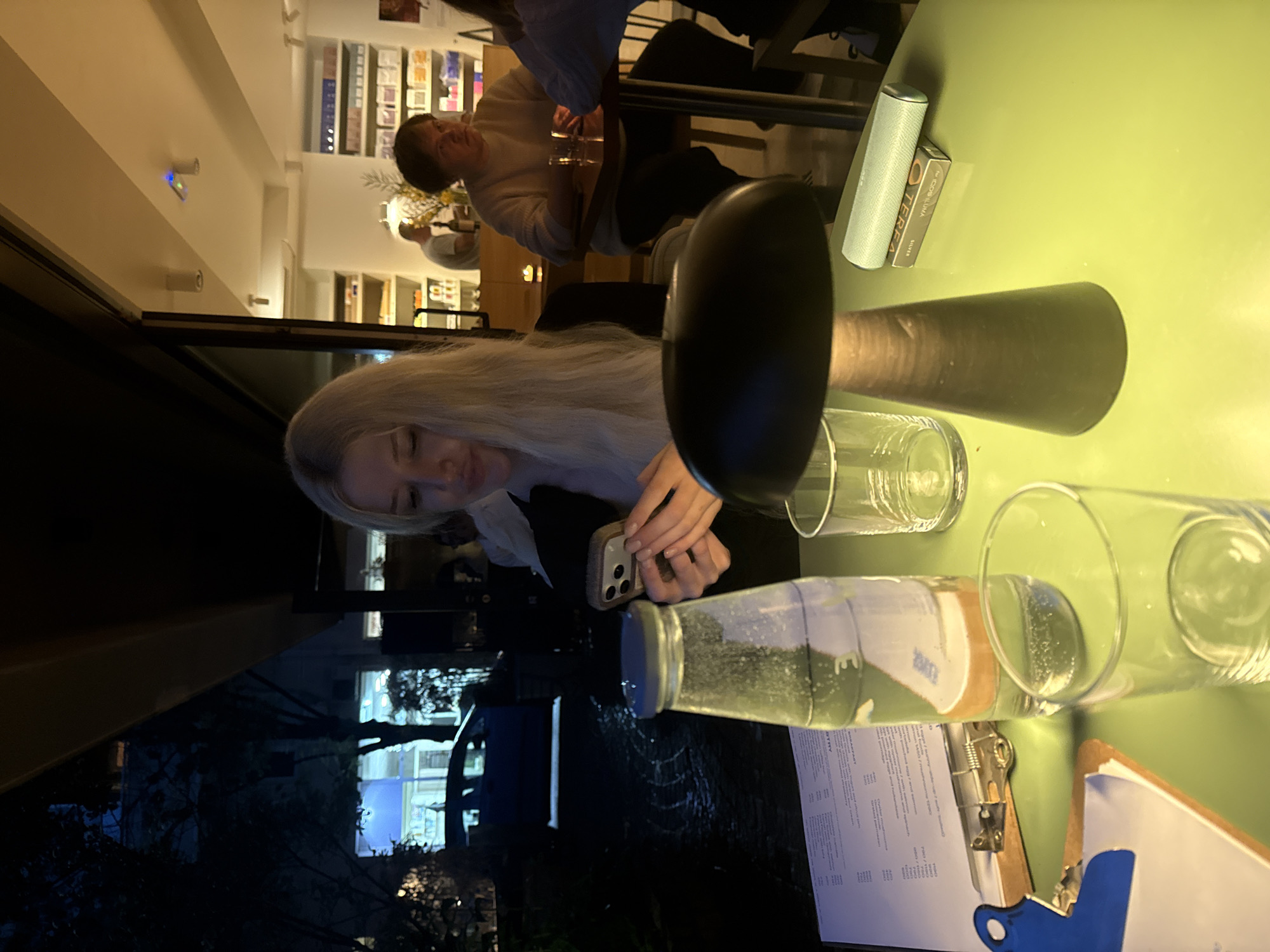 A rotated indoor shot shows a table with a young blonde woman using a smartphone. The table is set with glasses, a water bottle, a menu on a clipboard, a table lamp, and a small package. In the background, other patrons are visible, suggesting a restaurant setting. Through the window, urban scenery with bare trees and buildings can be glimpsed, likely in Belgrade, Serbia.