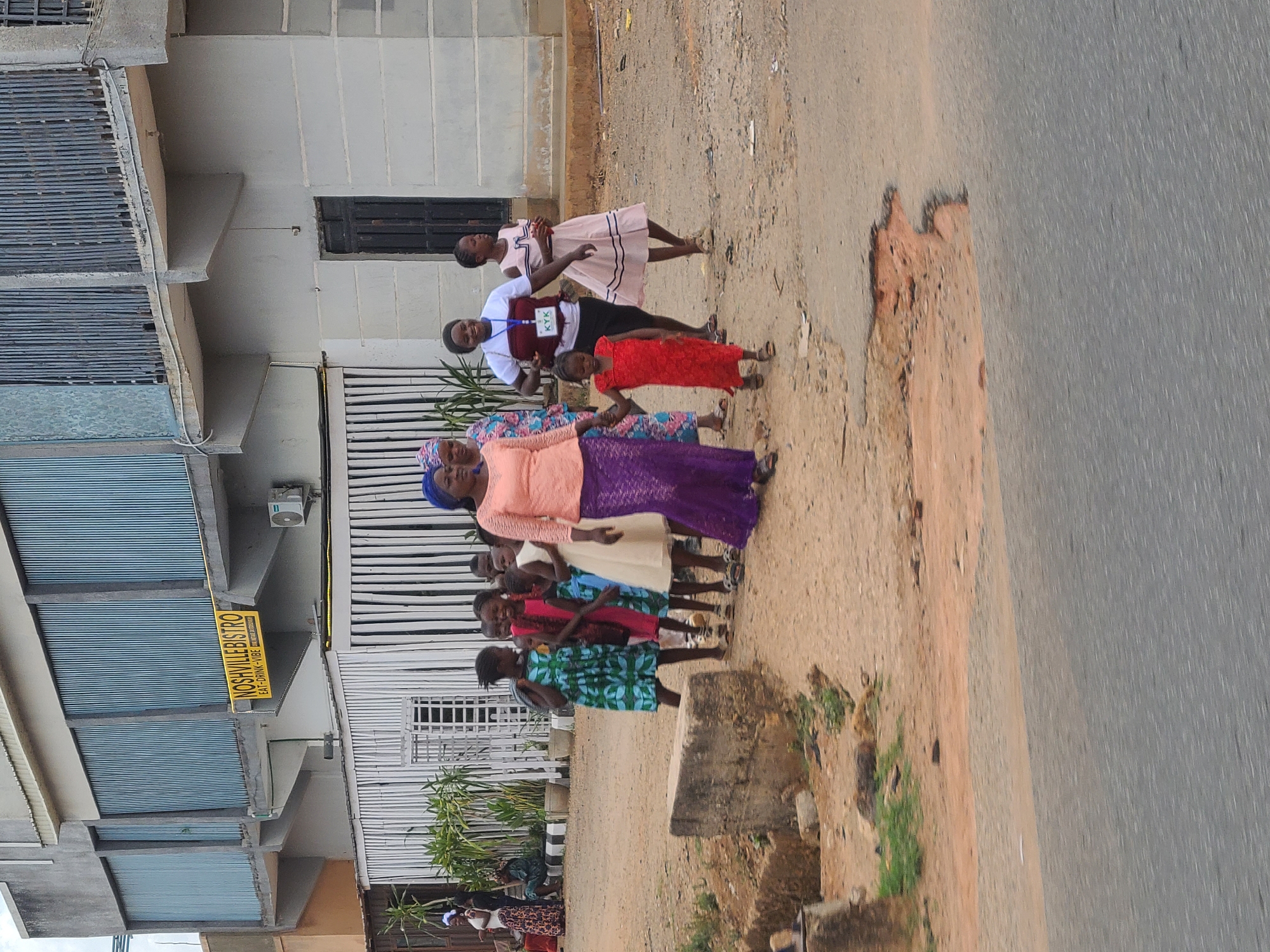 In Jos, Nigeria, a group of individuals, primarily children and young women, stand on a dirt area adjacent to a paved road. The individuals are diverse in appearance, with varying skin tones and attire. The clothing ranges from bright, patterned dresses to more subdued garments. One young woman wears a badge, indicating a possible role or affiliation. The surroundings include buildings and a sign that reads "Noshville Bistro Eat-Drink-Vibe". The environment appears to be a mix of urban and less developed areas.