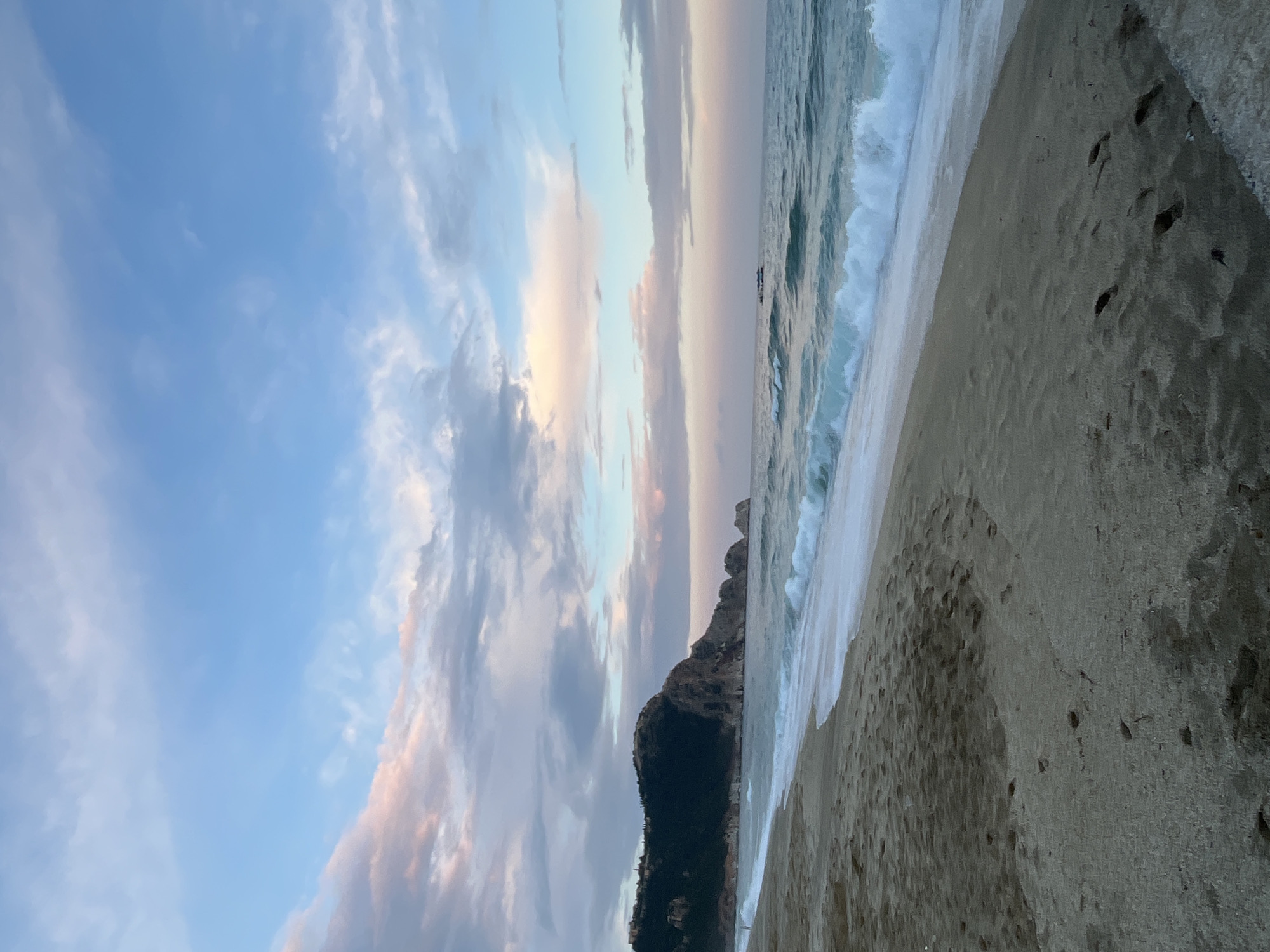 This image depicts a beach scene, likely in Alanya, Turkey. The foreground shows a sandy beach with clear footprints. The water's edge is marked by foamy waves crashing onto the shore. In the middle ground, a dark, rocky headland rises from the beach, extending into the sea. The background showcases a sky filled with pastel-colored clouds, predominantly blue and pink, suggesting either sunrise or sunset. The scene appears tranquil, with no visible people. The overall composition focuses on the natural beauty of the beach, coastline, and sky, with a focus on light and color.