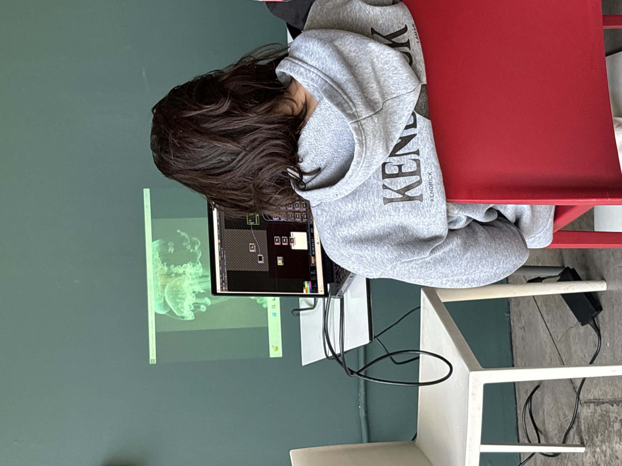 The image shows a person from behind, seated in a red chair and engrossed in a laptop. The individual has dark, wavy hair and is wearing a light grey hooded sweatshirt with the words "KENDRICK" and "LAMAR" printed in dark letters on the back. They are facing a dark teal-green wall.
The laptop screen displays what appears to be a visual programming or editing interface, characterized by a grid of small graphical modules, icons, and interconnecting lines. A timecode, "00:00:03:23", is visible, suggesting work on video, animation, or interactive media. To the left of the laptop, a bright, ethereal image of a jellyfish is projected onto the wall, likely related to the content on the screen.
A white table is partially visible to the person's right, and black cables with a power adapter are on the floor nearby. The scene depicts an indoor setting, possibly a studio, classroom, or dedicated workspace, emphasizing concentration on a creative or technical project. This activity takes place in Mexico City, Mexico.