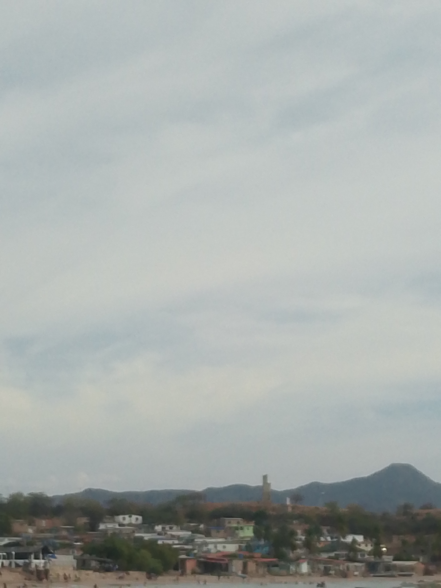 The image shows a distant view of Juan Griego, Venezuela, from the water. The foreground is blurred and indistinct, possibly water or sand. A densely packed town extends along the shoreline with small, brightly colored buildings interspersed with trees. Low-lying hills rise behind the town with a taller mountain visible in the distance on the right. A tall, narrow tower-like structure is visible in the middle ground, slightly left of center. The sky is overcast. The scene appears to be a populated beachfront.
