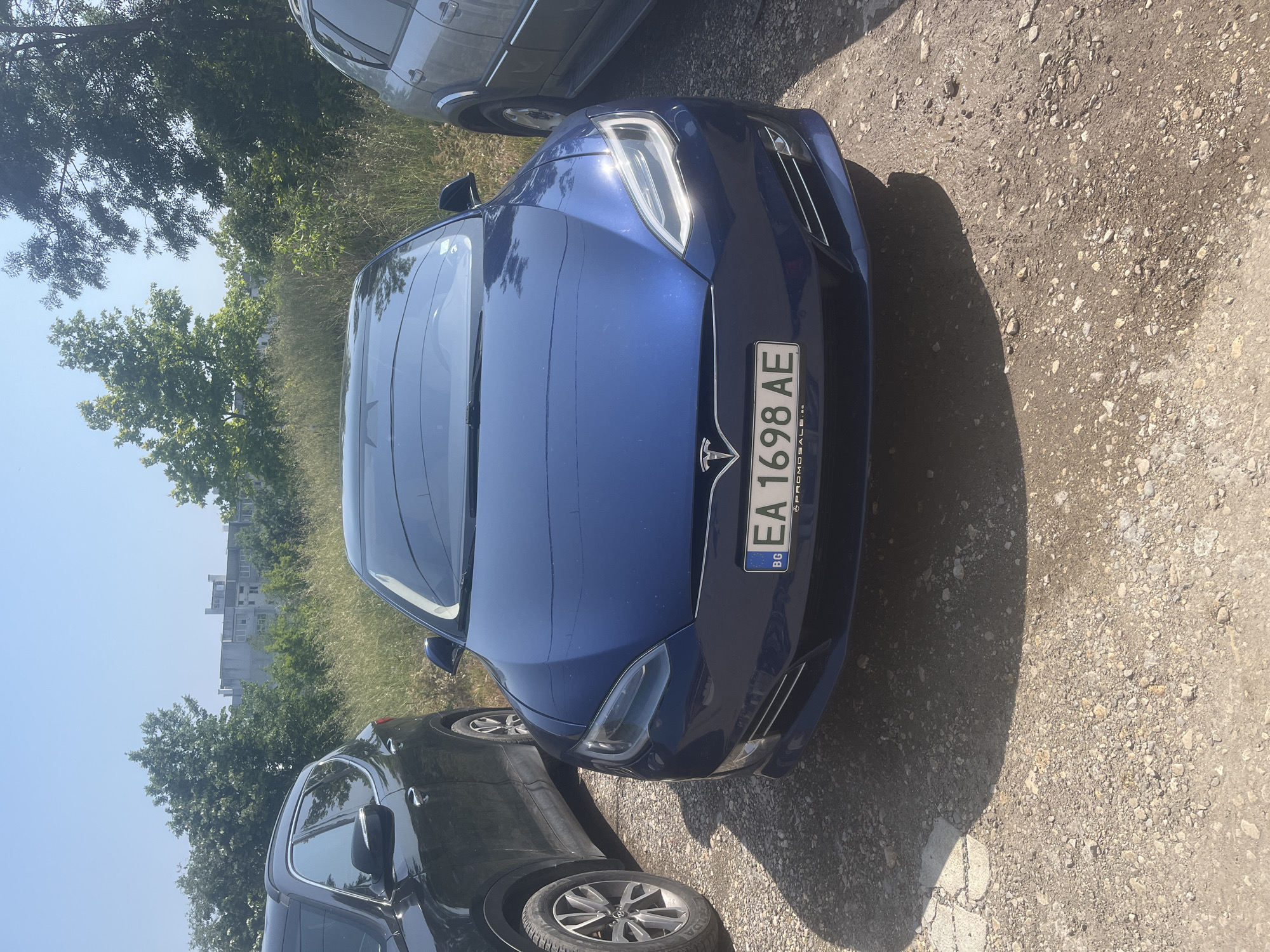 The image captures a blue Tesla Model S parked outdoors on a sunlit, unpaved surface primarily composed of dirt and gravel. The car is angled towards the viewer, with its front headlights illuminated and reflecting the bright sun. A Bulgarian license plate, featuring the country code "BG" and the registration "EA 1698 AE", is prominently displayed on the front bumper. Faint text below the number appears to be "PROMOSALE.eu".
The scene is set during a clear, sunny day, likely midday or early afternoon, as evidenced by the strong shadows cast on the ground, including a large, dark shadow extending from the right side. To the left of the Tesla, part of a gray SUV is visible, while on the right, a dark-colored SUV is also parked, with its front wheel, bearing a "FULDA" tire, clearly visible.
In the background, lush green trees and tall grass line the area, partially obscuring a distant, light-colored, multi-story building. The setting suggests an informal parking lot in Varna, Bulgaria, possibly on the edge of an urban area, characterized by its natural, unpaved ground and surrounding vegetation. No people are visible, and there is no specific activity taking place other than the vehicles being parked.