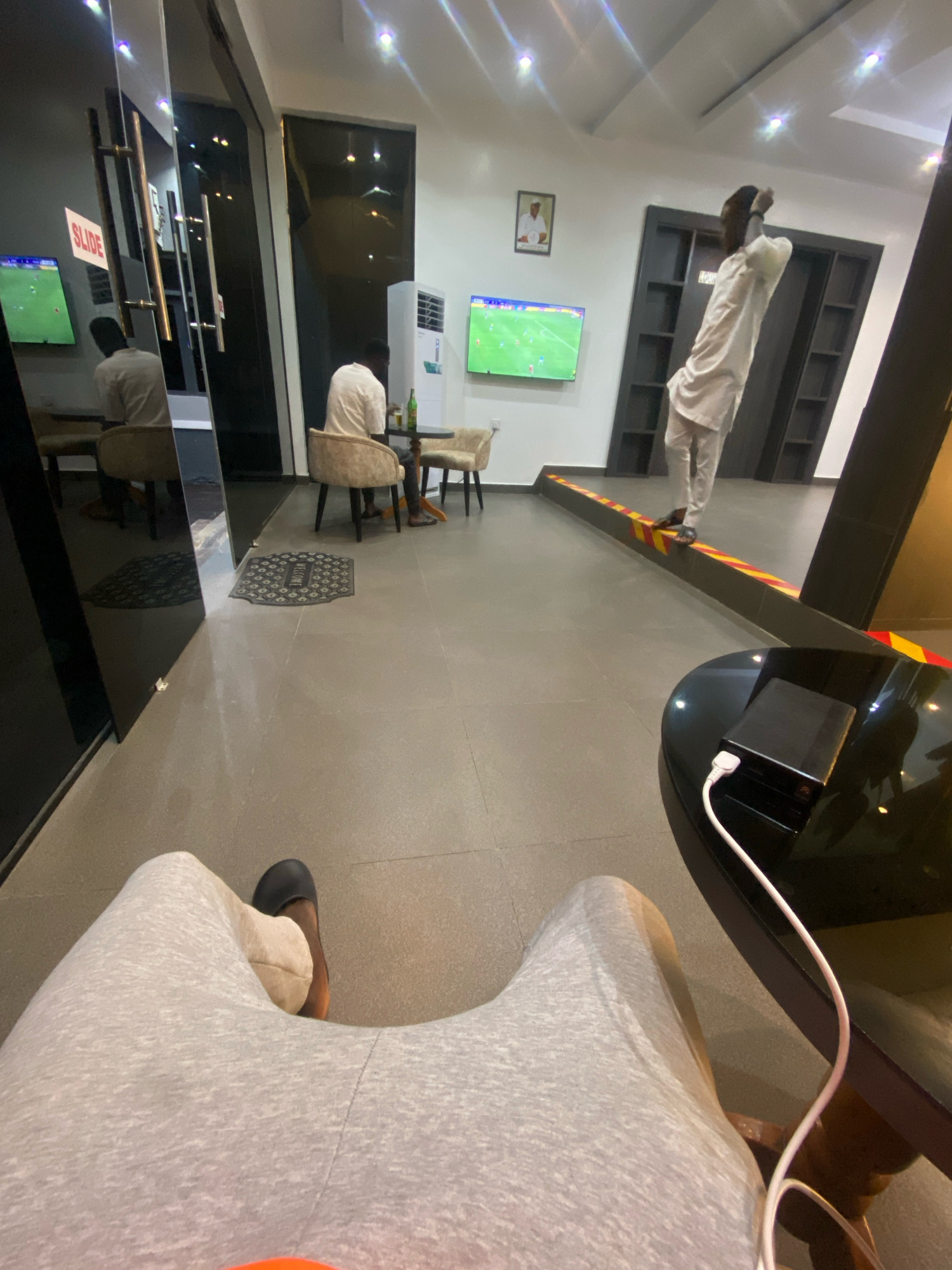 The image depicts a lounge area within a building, possibly a business or hospitality establishment, in Barden Yana, Nigeria. In the foreground, a person's legs are visible, clad in light gray sweatpants, with one foot wearing a dark slipper. The person appears to be seated, facing away from the camera.
In the mid-ground, two men are present. One man, wearing a white robe and pants, is standing and appears to be watching a television screen mounted on the wall. The television is displaying a soccer match. Another man, wearing a light-colored t-shirt, is seated at a small round table with chairs, holding a beverage.
The environment consists of a tiled floor, large reflective glass panels along one wall, and additional seating areas. A flat-screen television is also visible on another wall. A black round table with a power bank and a white cable is in the foreground on the right. The lighting appears to be artificial, with overhead lights illuminating the space. The overall ambiance suggests a relaxed setting where patrons can watch sports.