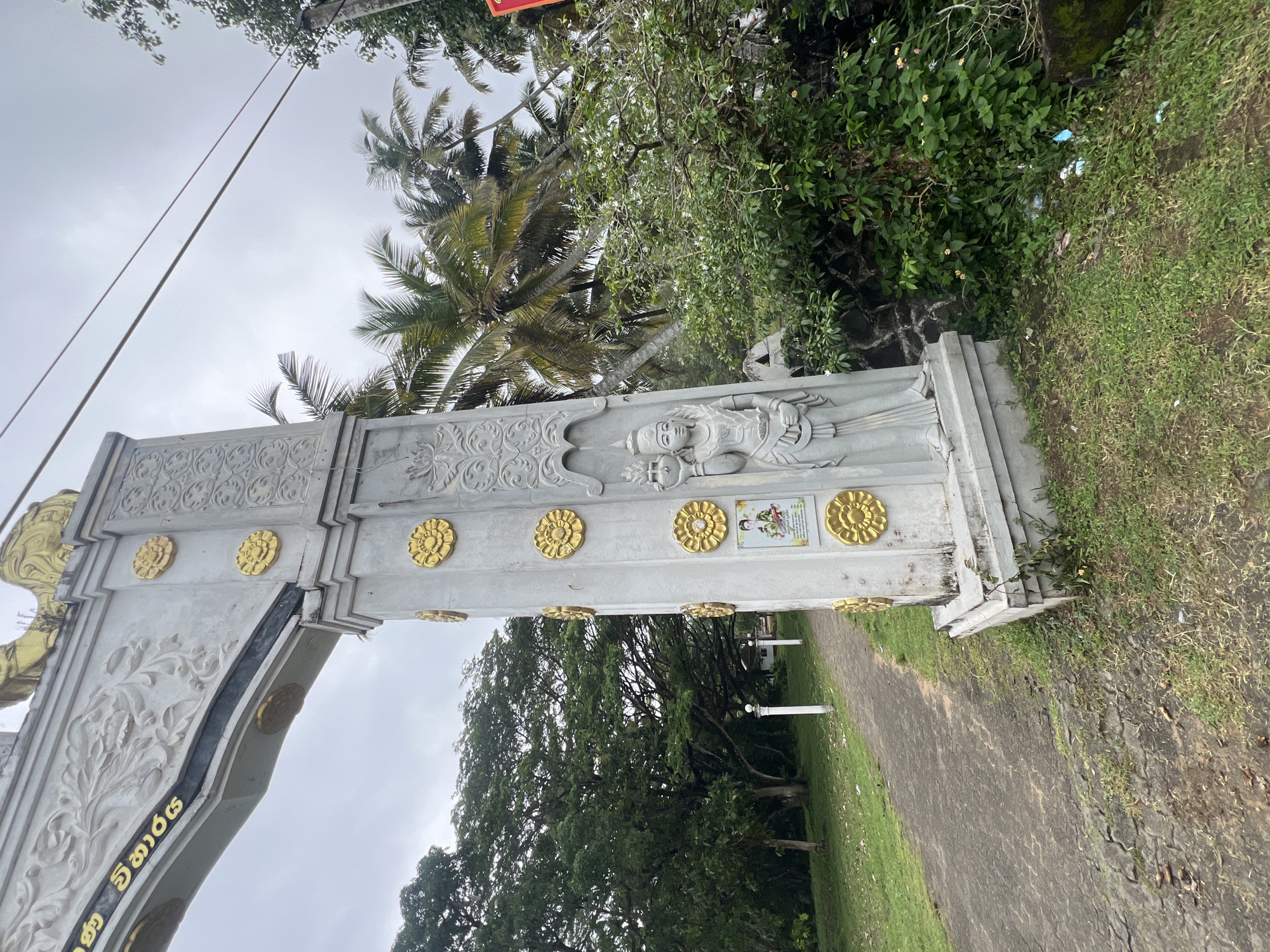A grey concrete archway gate stands in Boossa, Sri Lanka. The archway is decorated with intricate carvings and gold-colored floral motifs. A statue of a female figure, possibly a deity or historical personage, is carved into the pillar of the archway, holding a pot. Below the statue is a framed image. In the background, lush green trees, including palm trees, are visible under a cloudy sky. A paved pathway leads away from the archway, bordered by grass. The scene appears to be an entrance to a temple or a significant cultural site.