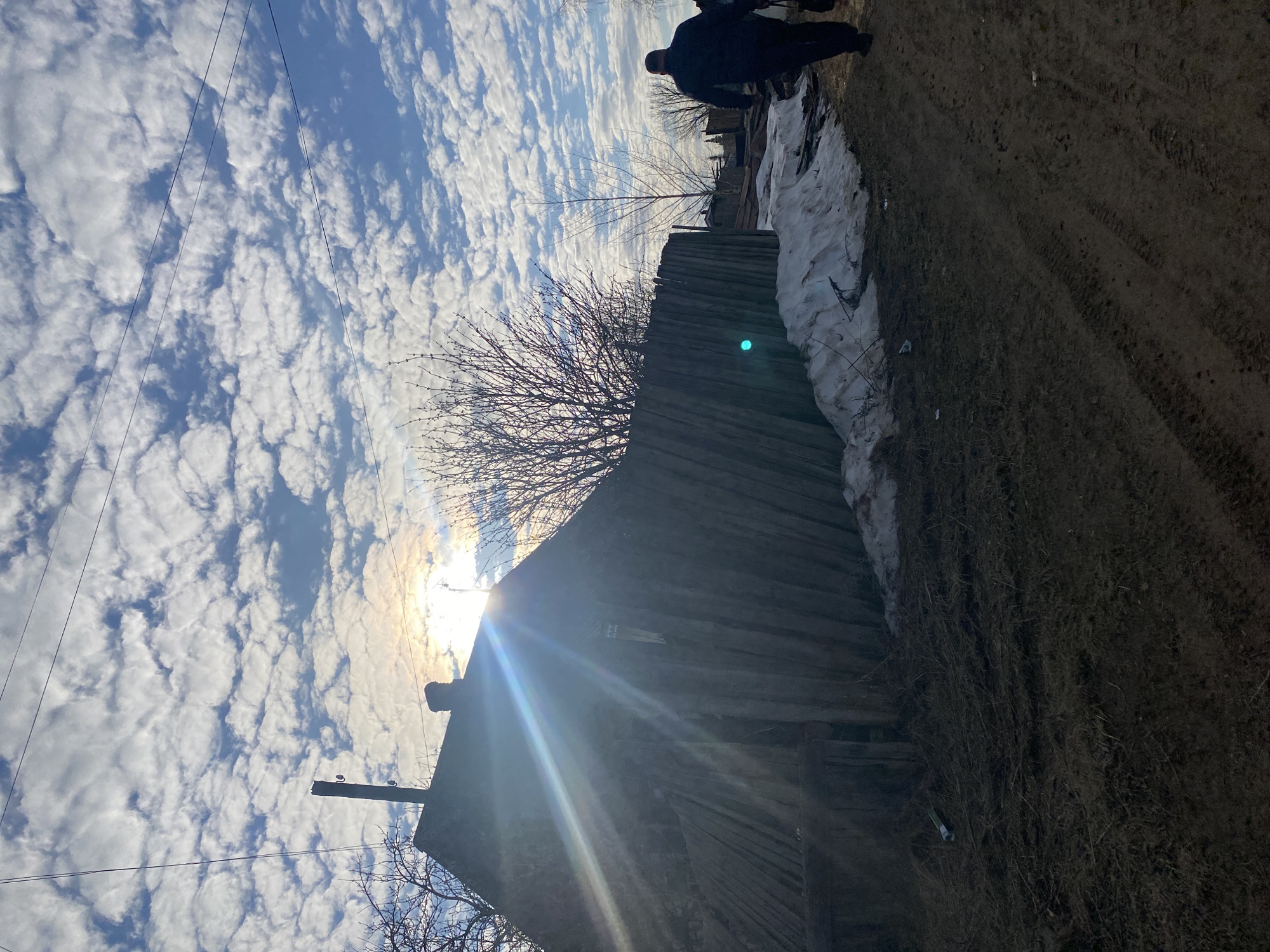 The image depicts a rural scene in Ольховка, Russia. The sky is filled with altocumulus clouds. A wooden structure, likely a shed or outbuilding, is prominently featured. A bare tree stands near the structure. Patches of snow remain on the ground. A dirt path or road is visible, and a silhouetted figure or figures are present in the background. A utility pole is visible with wires. The sun is positioned behind the structure, creating a lens flare effect.