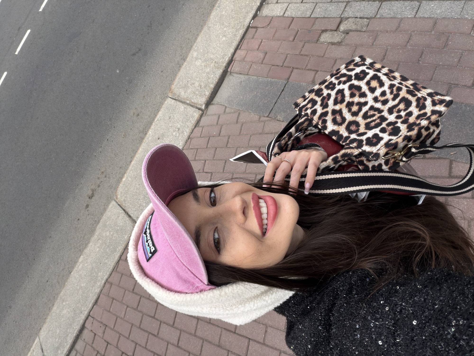 The image shows a woman posing for a selfie on what appears to be a city sidewalk. She is wearing a pink hat with a white fleece brim and a dark, sparkly sweater. Over her shoulder is a leopard-print bag with a striped strap. She has long brown hair and is wearing rings on her fingers. The background includes brick paving stones and a street with white lines, suggesting an urban environment. The bricks on the sidewalk are red and gray, and the lines on the street are painted white. The location is in the city of Saint Petersburg, Russia.