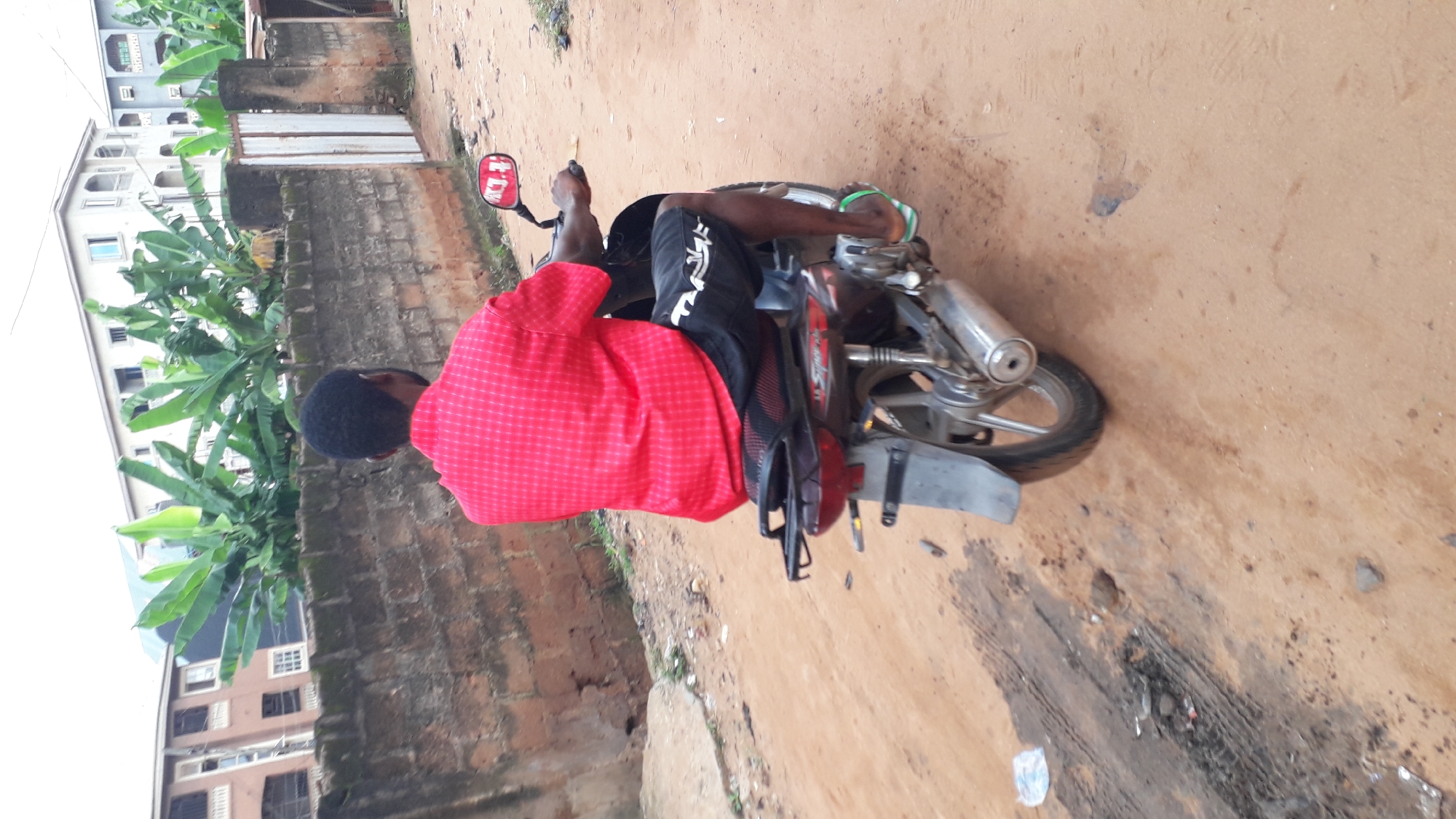 In Nkpor, Nigeria, a person sits on a red and black motorcycle parked on a dirt path next to a brick wall. The person, wearing a red checkered shirt and black shorts with white lettering, has their back to the camera and their head turned to the left. They are wearing flip-flops with green and white straps. The motorcycle has a silver exhaust pipe and a red rearview mirror. To the left of the motorcycle, there are large green banana leaves and a multi-story building with white and grey facades. The ground is made of loose brown dirt with some patches of dark, wet-looking earth. The scene appears to be outdoors during the day, with no indication of specific weather conditions. The overall impression is a snapshot of everyday life in a Nigerian town.