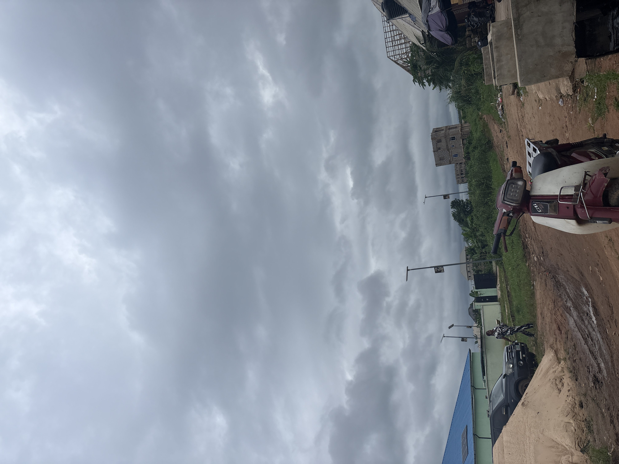 A wide shot captures an overcast sky above a street scene in Onitsha, Nigeria. In the foreground, a red and white motorcycle is parked on a dirt road. To the left, a pile of sand sits next to a black SUV and a person in a patterned outfit walks away from the camera. Behind them, a light green building with a blue roof is visible, and several streetlights dot the landscape. In the midground, unfinished buildings under construction stand amidst lush green vegetation. To the right, a small market stall with a dark umbrella is set up, and concrete structures line the foreground. The overall impression is one of a developing urban area on a cloudy day.
