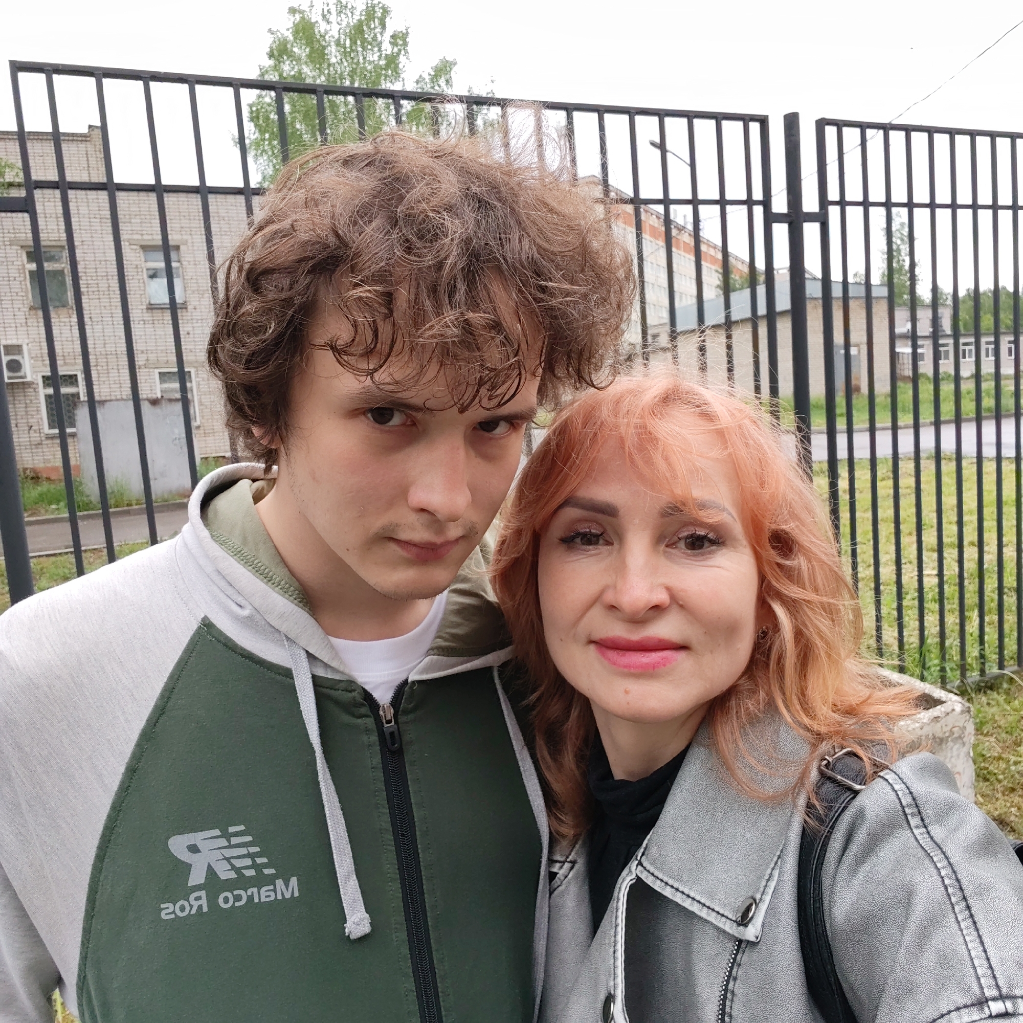 A medium shot captures a young man and a woman standing close together in an outdoor setting. The man, on the left, has brown curly hair, dark eyes, and fair skin. He wears a grey and green hooded sweatshirt with a white t-shirt underneath. The brand logo "Marco ROS" is visible on the sweatshirt's chest.
The woman, on the right, has light reddish-orange hair, and light skin. She wears a shiny silver-grey jacket over a black turtleneck. A black strap of a bag hangs from her shoulder.
Behind them is a black metal fence. Behind the fence are building structures, with some green foliage visible in the background. The location is identified as Chaikovsky, Russia. The image suggests an overcast day.