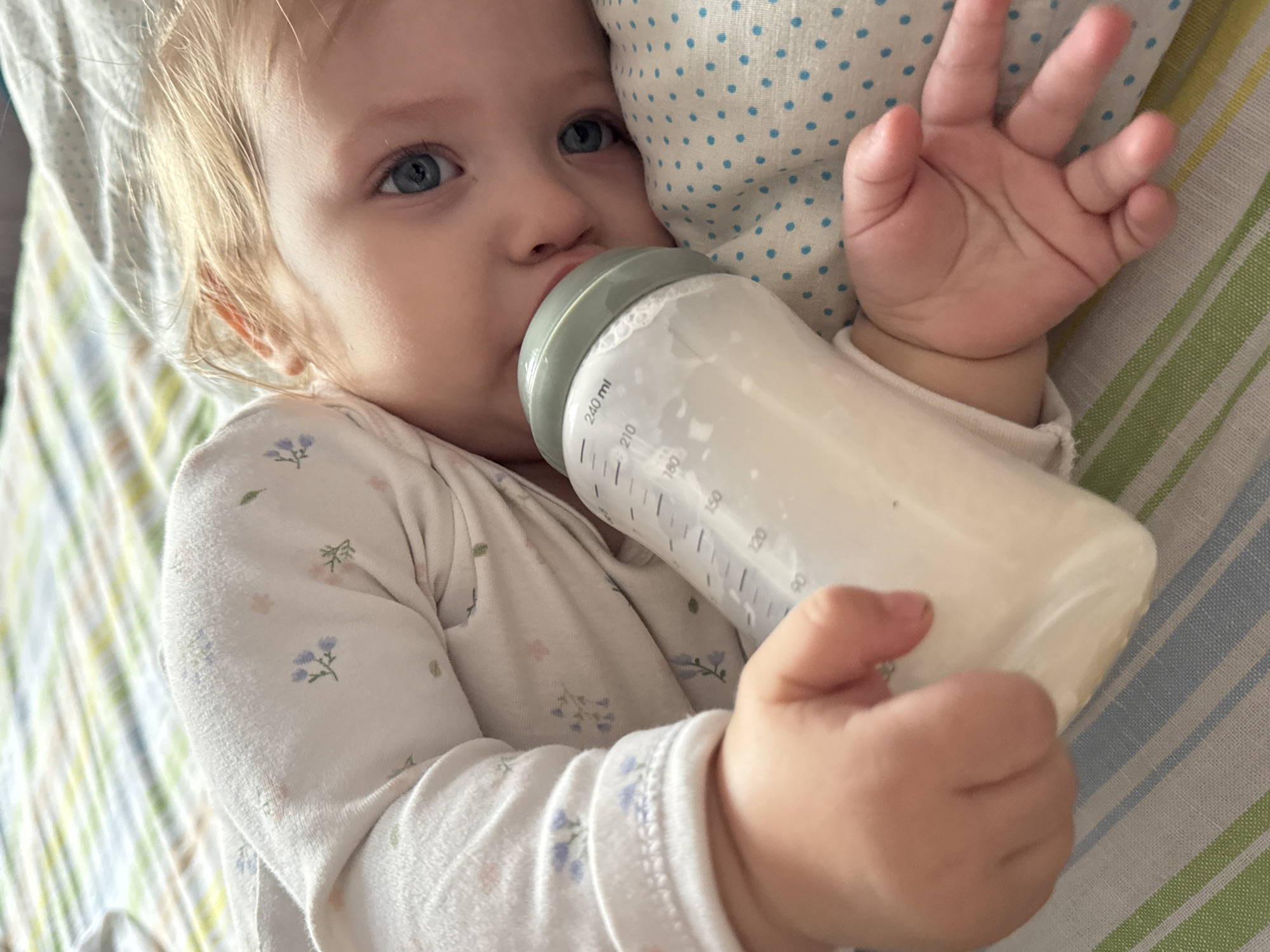 A baby with light skin and blue eyes is lying down and drinking from a baby bottle. The baby is wearing a long-sleeved white shirt with a floral print. The bottle is clear with a gray top and has markings in milliliters. The baby is holding the bottle with both hands. The baby is lying on a striped blanket and resting against a polka-dotted pillow.
The location is Nizhnyaya Gar', Russia.