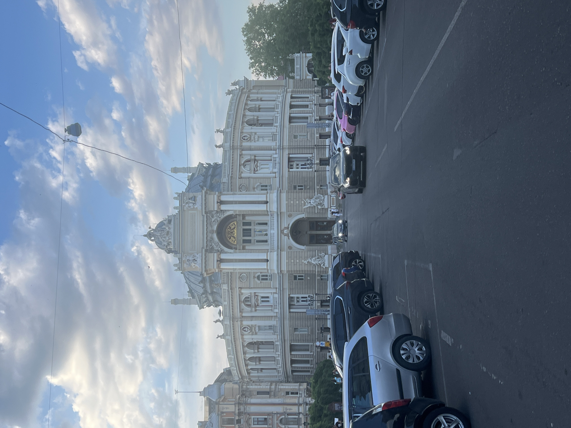 The image shows the Odesa Opera and Ballet Theater in Odesa, Ukraine. The grand, semi-circular building, constructed in a Baroque style, dominates the background. The facade is ornate, featuring columns, statues, and a prominent dome. In front of the theater, a parking lot is filled with various cars. The foreground and midground are occupied by these vehicles, with several black SUVs, a silver station wagon, and other cars parked in designated spots. The road surface is asphalt. A few people are visible walking in the distance near the theater entrance and around the parking area. The sky above is filled with dramatic clouds, suggesting either dawn or dusk. A lamppost hangs suspended by wires across the sky.