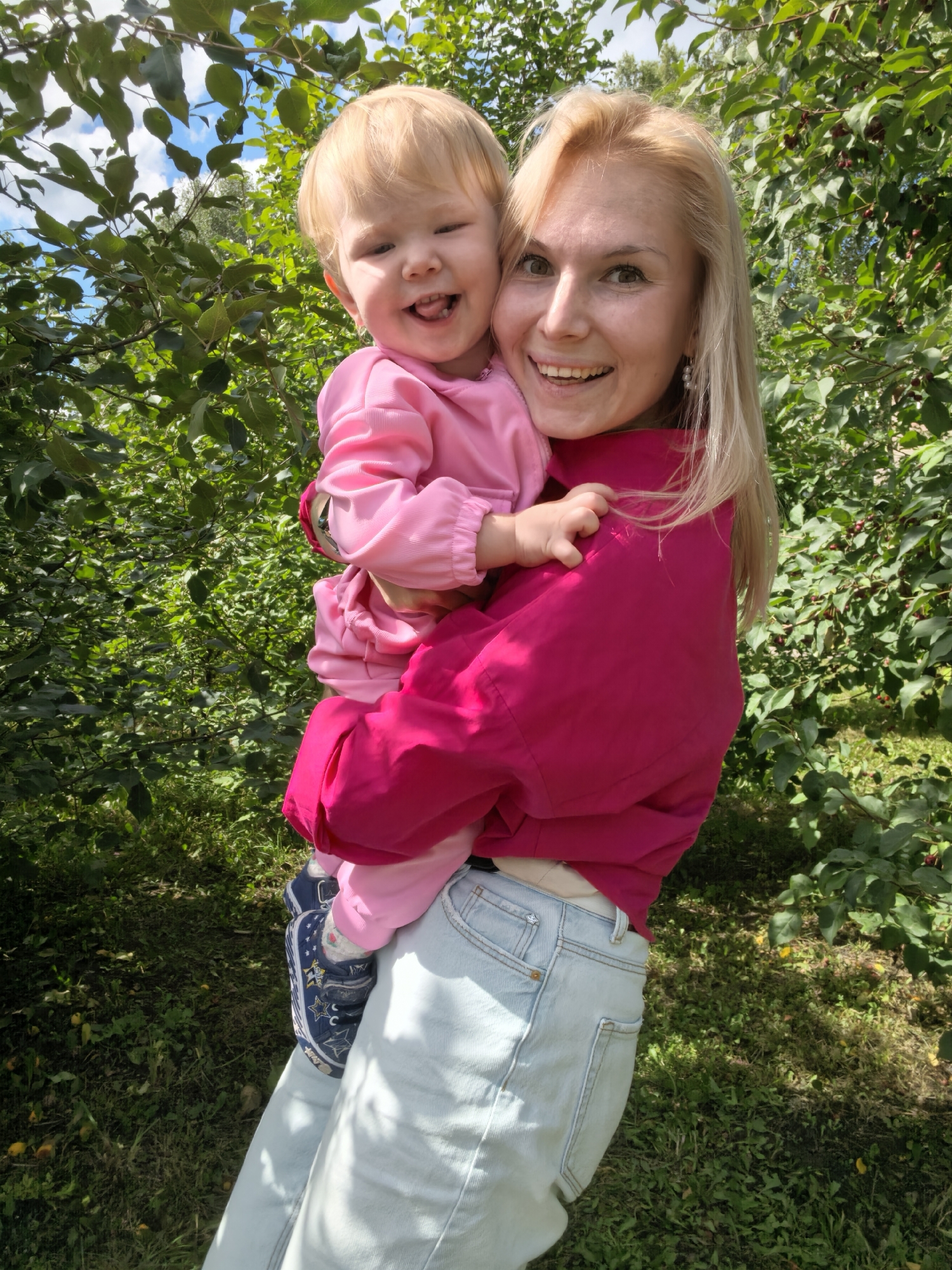 In Sterlitamak, Russia, a blonde woman in a bright pink shirt and light blue jeans holds a blonde toddler wearing a pink outfit. The child is smiling with its tongue out. They are standing outdoors amongst trees with green leaves and some red berries. The ground is covered in grass and fallen yellow leaves. The lighting suggests it is daytime.