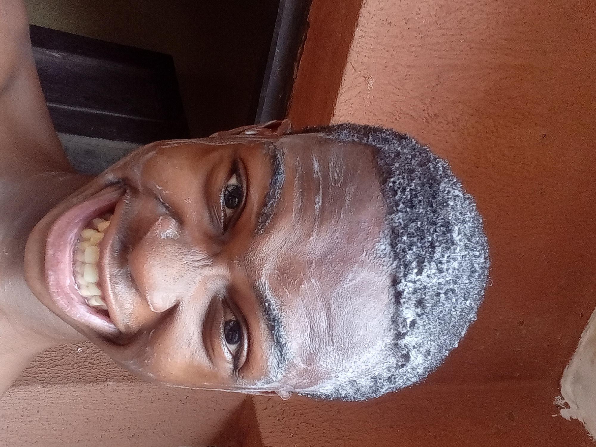 A man's face fills most of the frame. He has dark skin, close-cropped dark hair with what appears to be white foam covering the top of his head and face. He is smiling widely, revealing his teeth. The background includes a dark brown wall and what appears to be a dark-colored door. The location is Onitsha, Nigeria. The image is taken from a low angle, looking up at the man.