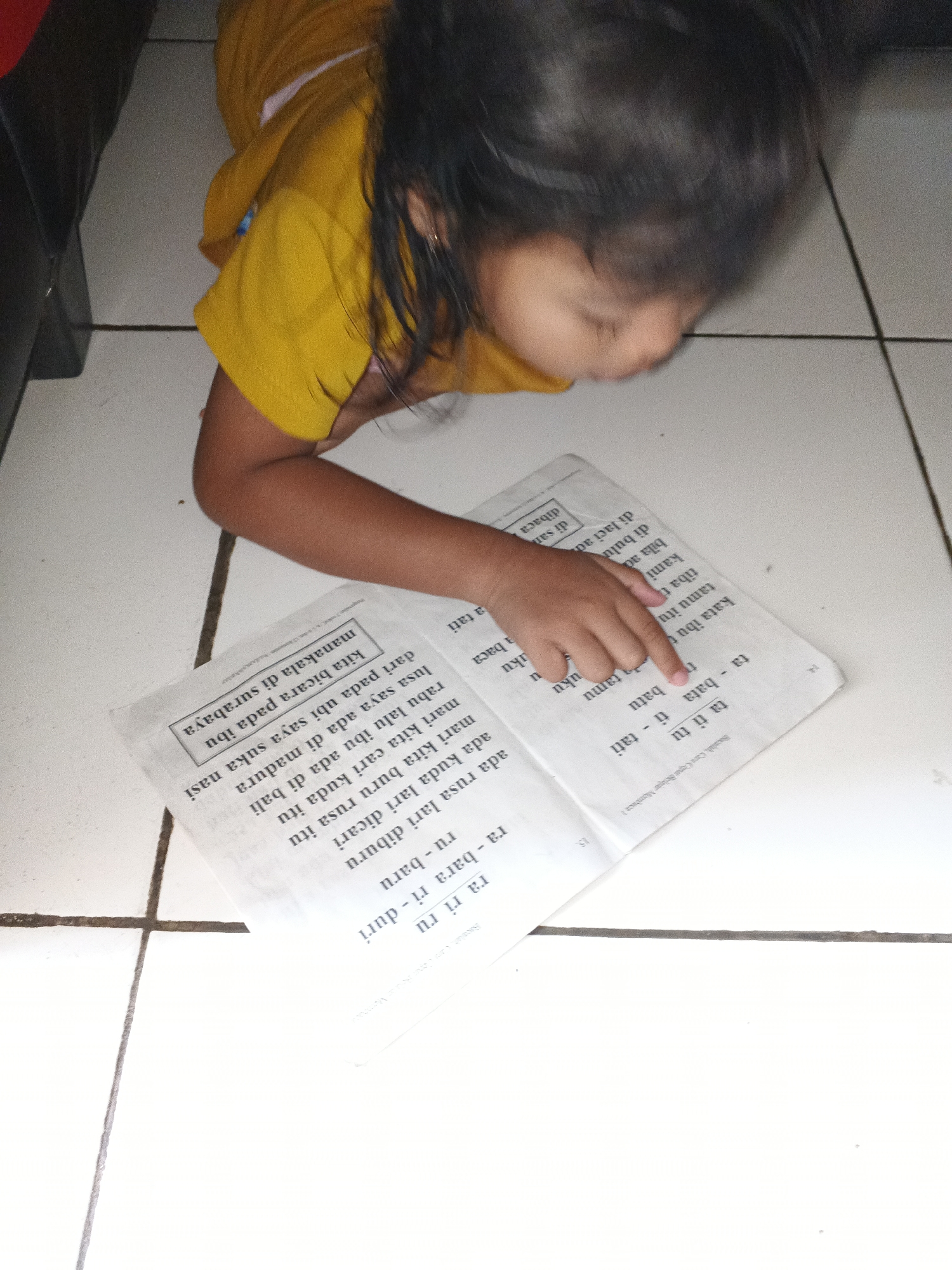 A young girl with dark hair is sitting on a tiled floor, wearing a yellow shirt. She is looking down at an open book and pointing at the text with her right index finger. The book has several lines of Indonesian text, and some words are highlighted or boxed. The floor appears to be made of white tiles with visible grout lines. The lighting suggests an indoor setting, possibly a home in Warungkiara, Indonesia, where the girl is engaged in reading or learning.