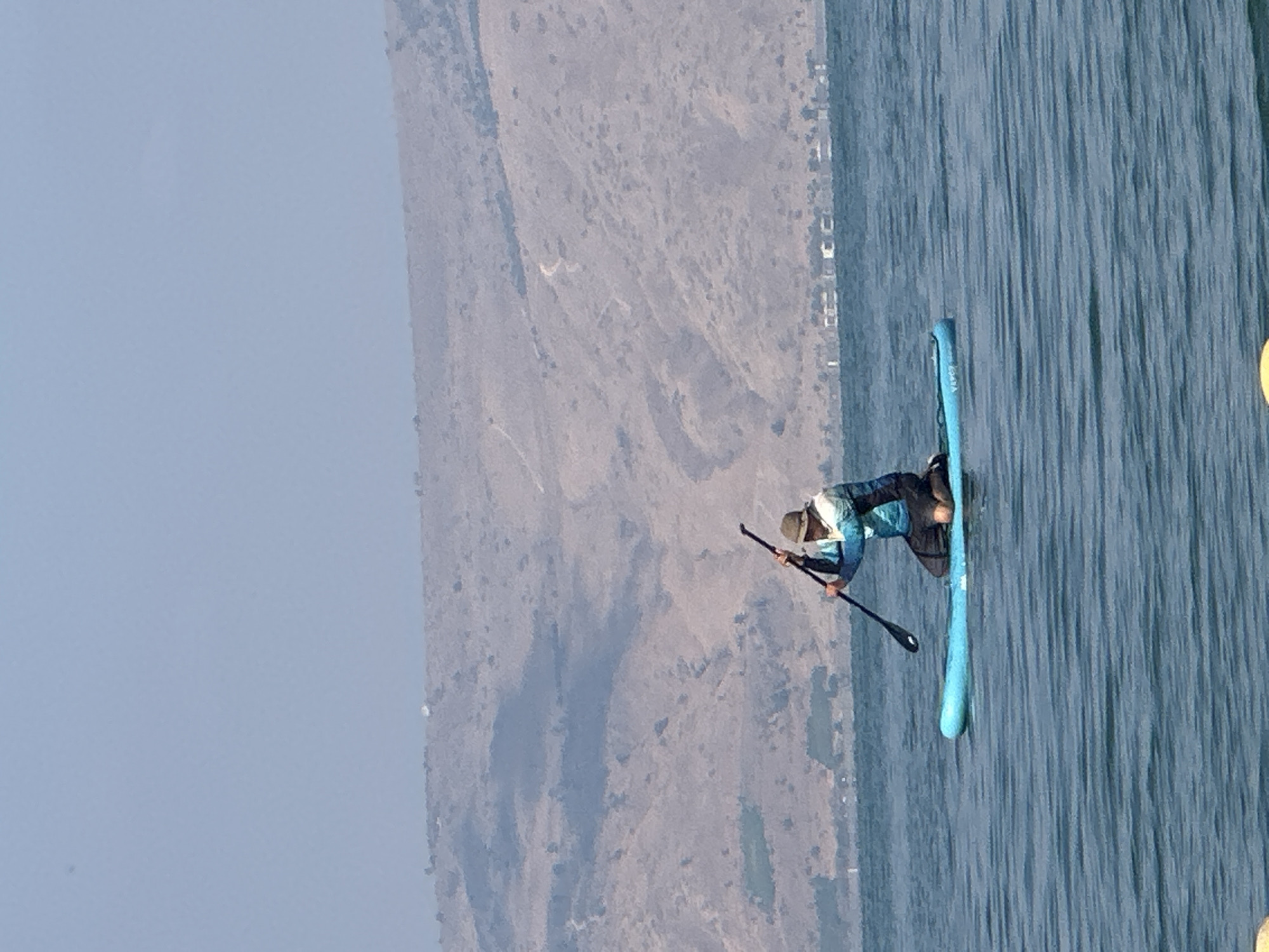 A person is paddleboarding on the Sea of Galilee in Tiberias, Israel. The individual is kneeling on a blue paddleboard, holding a paddle. The person is wearing a hat and a blue and white patterned jacket. The background features a dry, mountainous landscape with a clear blue sky. The water is choppy with visible ripples.