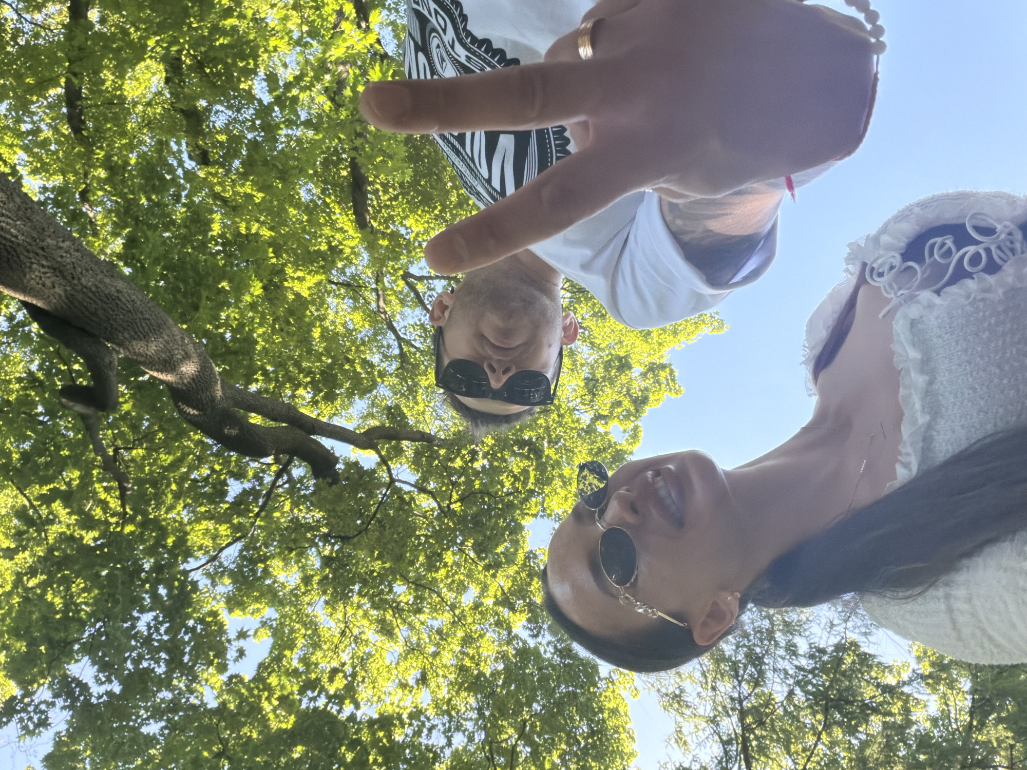 An outdoor, low-angle photograph taken in Saint Petersburg, Russia, captures two individuals looking up towards the camera under a canopy of green trees and a clear blue sky.
In the lower-left of the frame, a woman is visible, wearing dark oval sunglasses with decorative gold-toned frames and a light-colored, possibly white or cream, textured top with ruffled trim. Her dark hair is pulled back in a ponytail, and a subtle smile is apparent beneath her sunglasses. She also wears a delicate necklace and an earring on her visible ear.
Positioned in the upper-right, slightly behind and above the woman, a man is seen wearing dark rectangular sunglasses and a white short-sleeved t-shirt featuring a distinctive black and white patterned graphic on the upper chest/shoulder area. His visible forearm displays tattoos. His right hand is prominently featured in the foreground, making a 'V' peace sign. A gold-colored ring is visible on his right ring finger, and a white beaded bracelet is on his wrist.
The background consists of a dense canopy of vibrant green leaves and tree branches, with several sections of bright blue sky visible through the foliage. The scene is brightly lit by natural sunlight, suggesting a clear day in an outdoor, heavily treed environment such as a park or garden within Saint Petersburg.