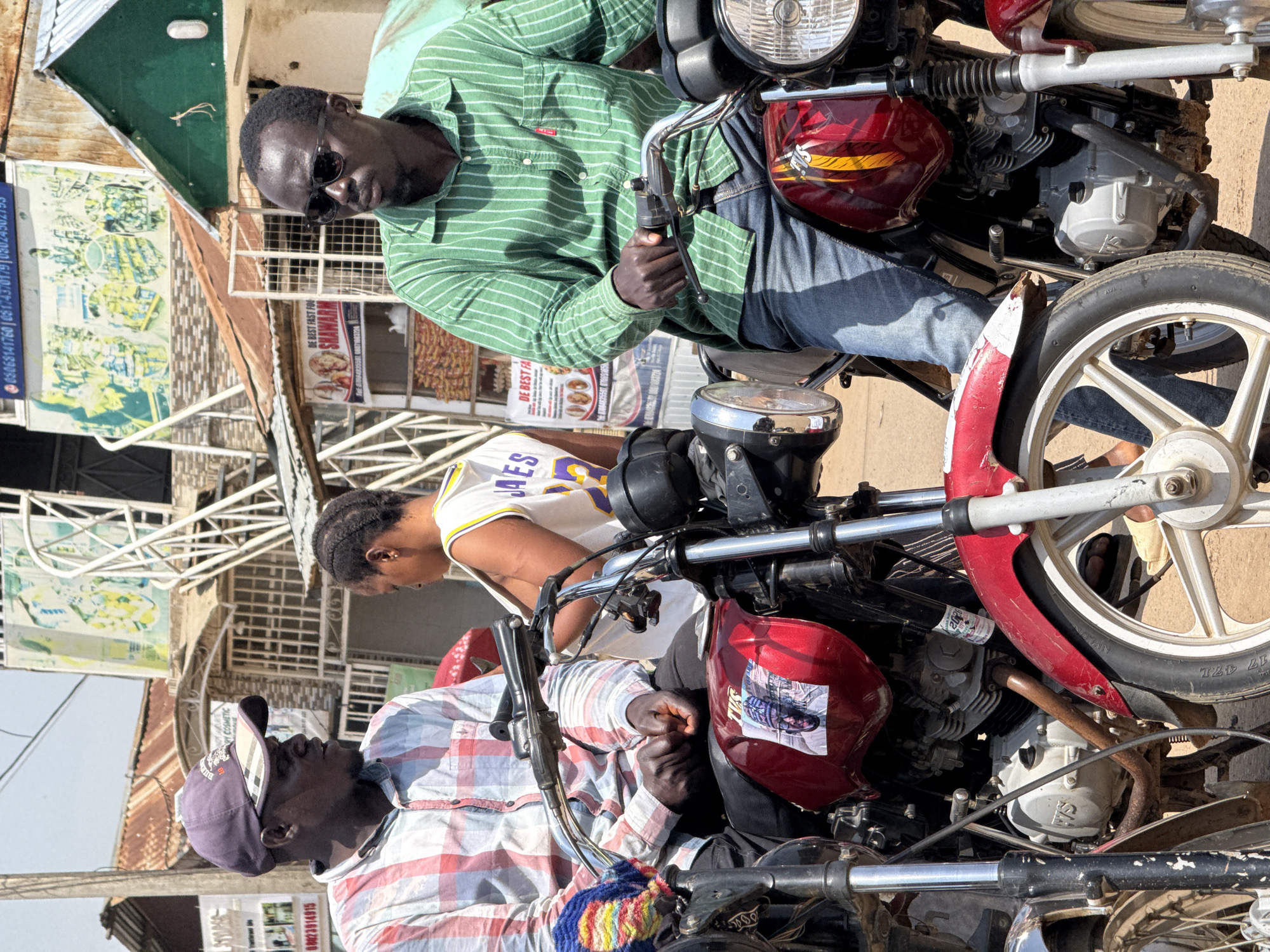 In the city of Abuja, Nigeria, three individuals are positioned on or near two motorcycles. The central figure, a person with braided hair wearing a white and yellow tank top, is seated between the two motorcycles. To the left, a man wearing a red and white plaid shirt and a purple baseball cap is seated on a motorcycle. To the right, a man in a green striped shirt and sunglasses is also seated on a motorcycle. Both motorcycles are red and appear to be the same make and model. The background reveals a street scene with buildings, signage, and what appears to be market stalls. The overall lighting suggests it is daytime.