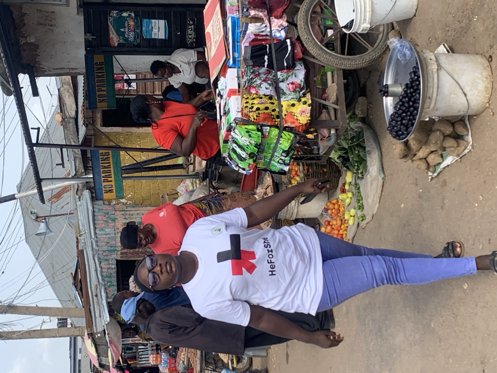 The image shows a woman walking in a busy marketplace in Jos, Nigeria. She is wearing a white t-shirt with the "HeForShe" logo and blue pants. She is looking directly at the camera with glasses on. Behind her, vendors are selling produce and other goods from stalls and carts. The market appears to be outdoors, with corrugated metal roofs and various structures visible. The ground is dirt and uneven. The overall atmosphere is bustling and vibrant.