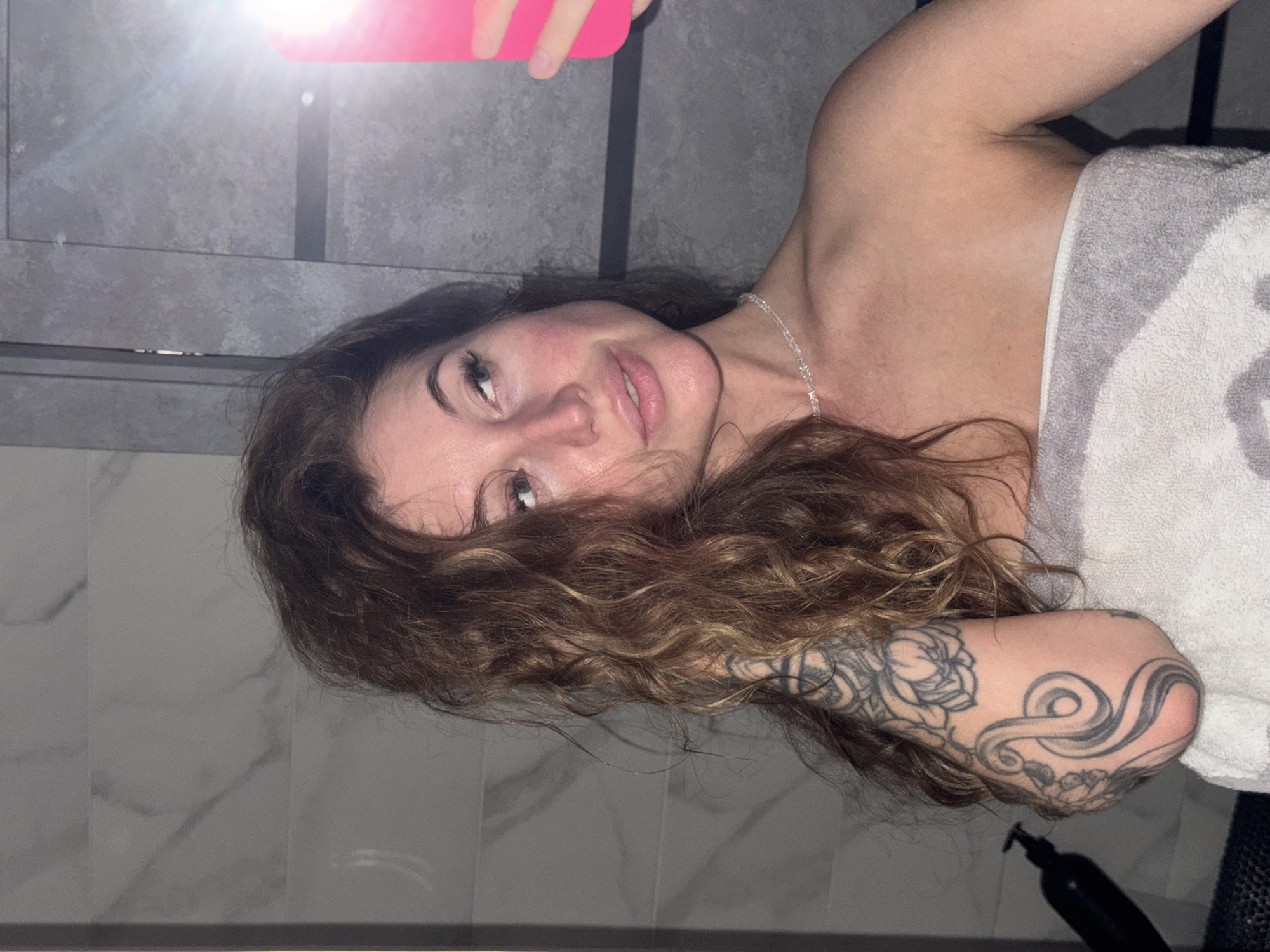 A woman with curly brown hair, a silver necklace, and a tattoo on her left arm is taking a selfie in a bathroom with light grey marble-like tiles. She is partially covered by a grey and white towel. A black bottle sits on the ledge near her arm. A pink smartphone is visible in the upper left corner of the frame. There is no specific information that places this scene in Chaikovsky, Russia.