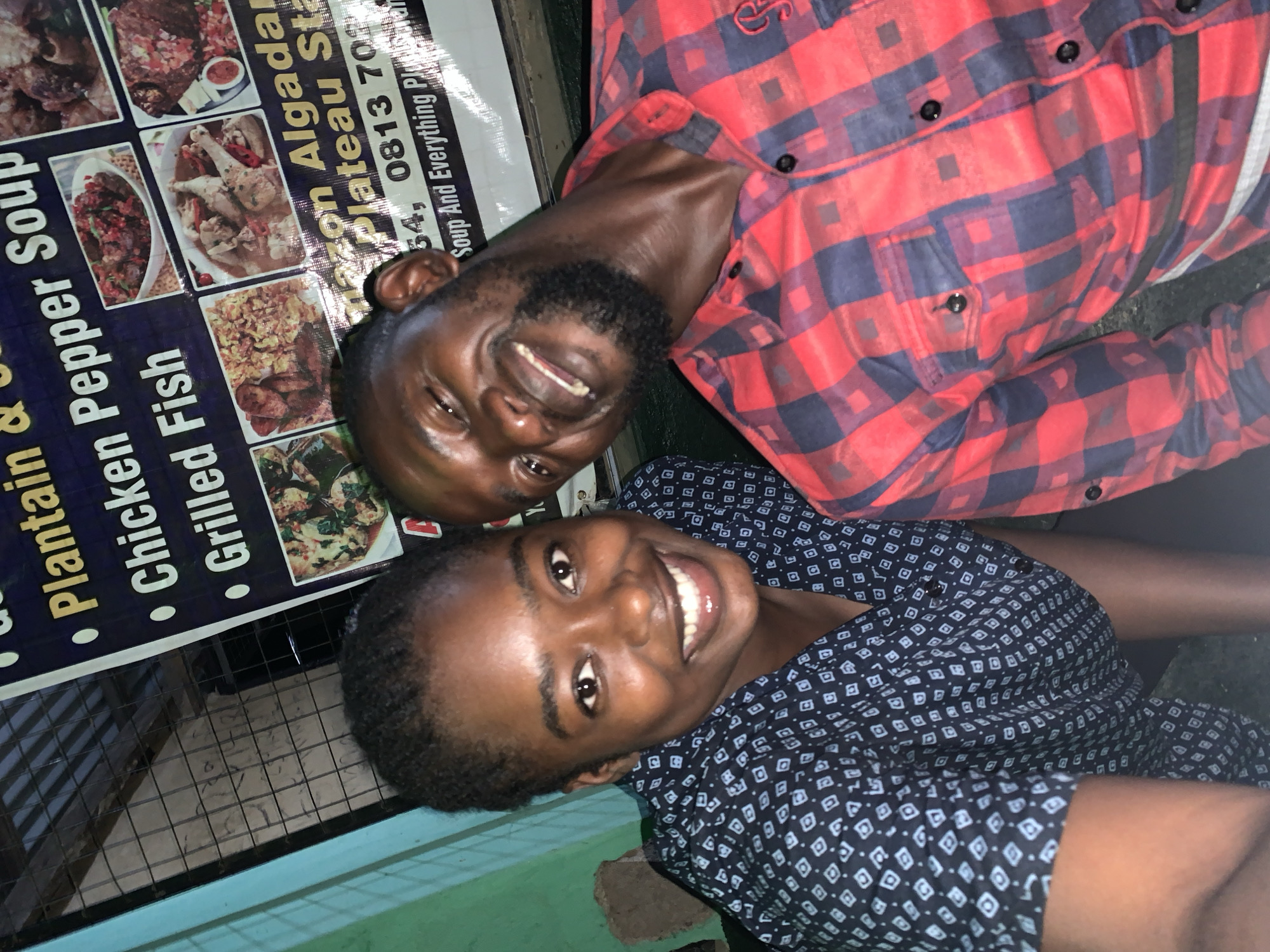 Two smiling dark-skinned people, a man and a woman, are positioned close together. Behind them is a sign advertising "Plantain & Pepper Soup" and other dishes, along with the address "Nazon Algada," which is in New Stadium, Plateau State, Nigeria, as well as the phone number "0813 701." The man is wearing a red and navy blue plaid shirt, while the woman is wearing a dark patterned shirt. The overall lighting in the scene is low.