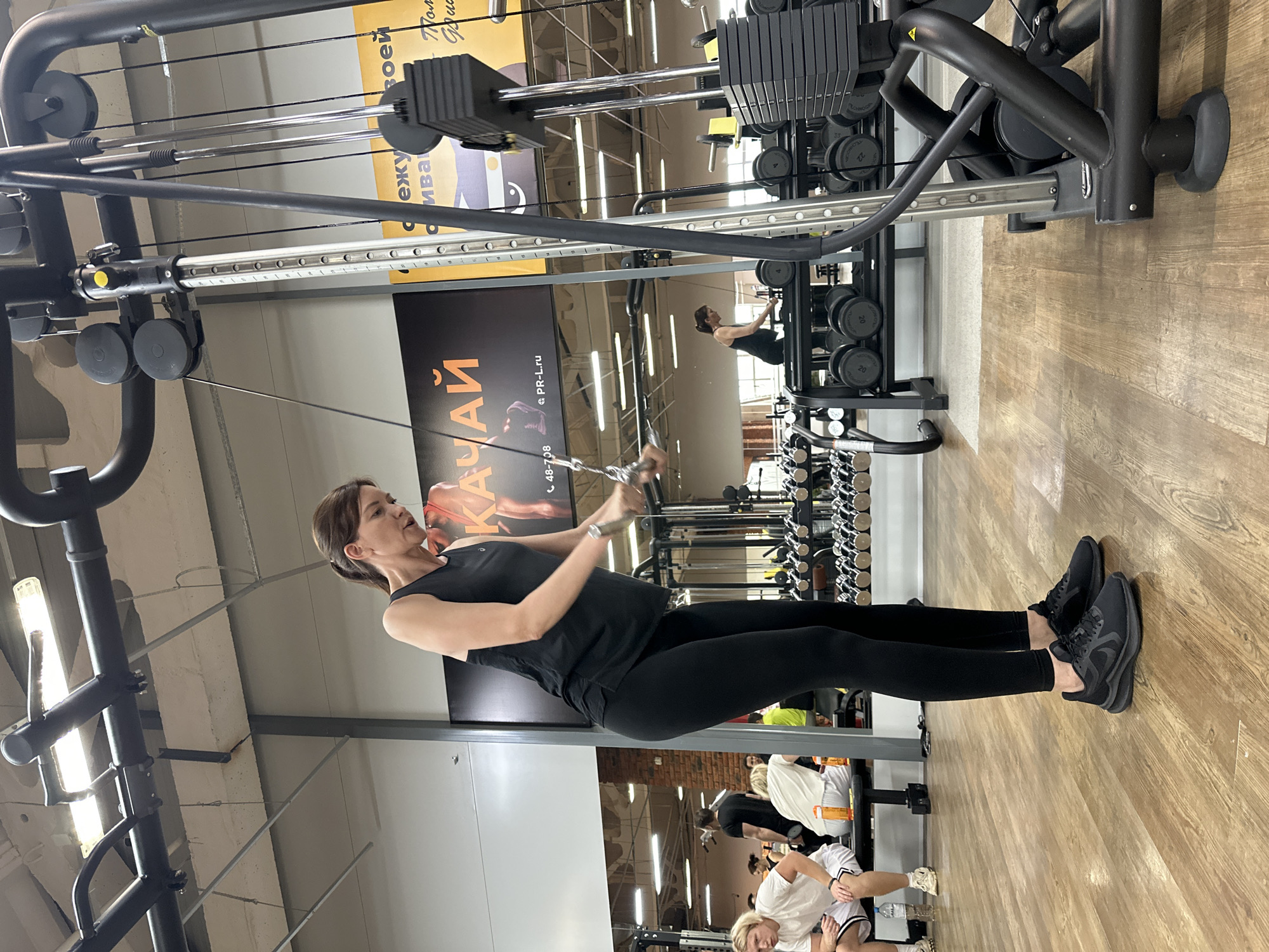 The image depicts a woman in a gym setting, specifically the Olympic gym in Chaikovsky, Russia. She is performing a cable pull-down exercise, dressed in a black tank top and leggings, wearing black athletic shoes. The exercise machine is black and features weight stacks. In the background, other gym equipment and individuals are visible, including racks of dumbbells, exercise benches, and other gym patrons. The gym has wooden flooring and overhead lighting. A promotional banner is visible in the background with text in Cyrillic and the website address "PR-L.ru".