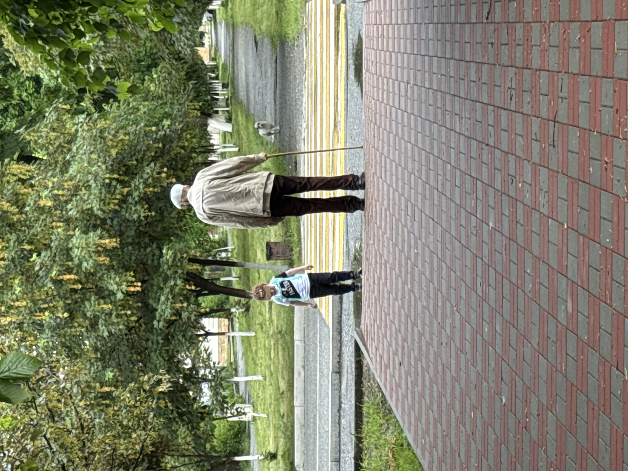 The image captures a scene in the city of Karachayevsk, Russia, featuring a pedestrian crossing with two individuals and a dog. In the foreground, a child, wearing a cap, t-shirt with the word "FASTER" printed on it, and black pants, stands facing away from the camera and looking down. To the right, an elderly man, dressed in a light-colored jacket, brown pants, and a cap, stands with his back to the camera, holding a walking stick. He is also facing away from the viewer, and appears to be looking in the same direction as the child. A light-colored dog is visible further down the pathway, facing the man and child. The setting is a park or a tree-lined avenue, with a paved walkway in the foreground and a pedestrian crossing with yellow and white stripes in the middle ground. Trees with green foliage and some yellow, hanging blossoms line the pathway on both sides. The ground is a mixture of paved brick and grass, with some puddles visible on the walkway near the crossing, indicating recent rain. The overall impression is a calm, everyday moment in a public outdoor space.