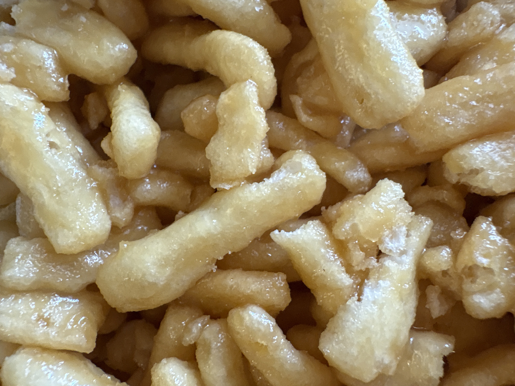 The image shows a close-up of a pile of Chak-chak, a Turkic dessert, with no discernible connection to the city of Chaikovsky, Russia. The Chak-chak pieces are irregularly shaped, lightly golden-brown, and coated with a glistening, translucent syrup. The texture appears slightly rough and uneven. The image is well-lit, with the light reflecting off the syrupy coating. No other objects or details are visible.