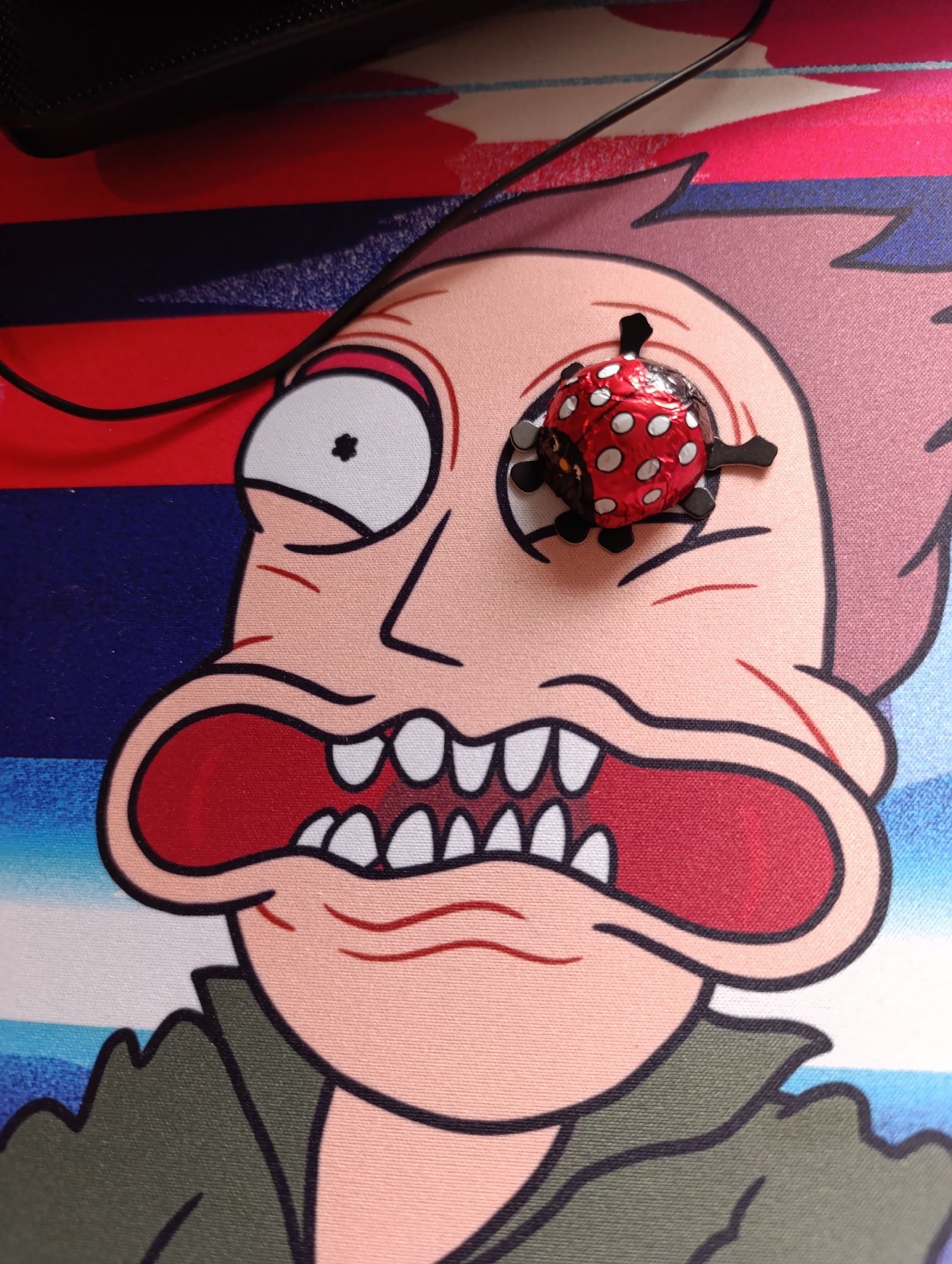 A close-up image shows a cartoon character printed on what appears to be a mousepad or similar surface. The character has exaggerated features, including a wide open mouth with numerous teeth, a large white eye with a dark pupil, and skin that is peach-colored. The background consists of horizontal stripes in red, blue, and white. A small, round chocolate wrapped in red foil with white polka dots, shaped like a ladybug, is placed over the character's other eye. The overall setting appears to be indoors, likely on a desk, as evidenced by a portion of what seems to be a black electronic device visible at the top of the frame. There is no information in the image that indicates the location is in Sofia, Bulgaria.