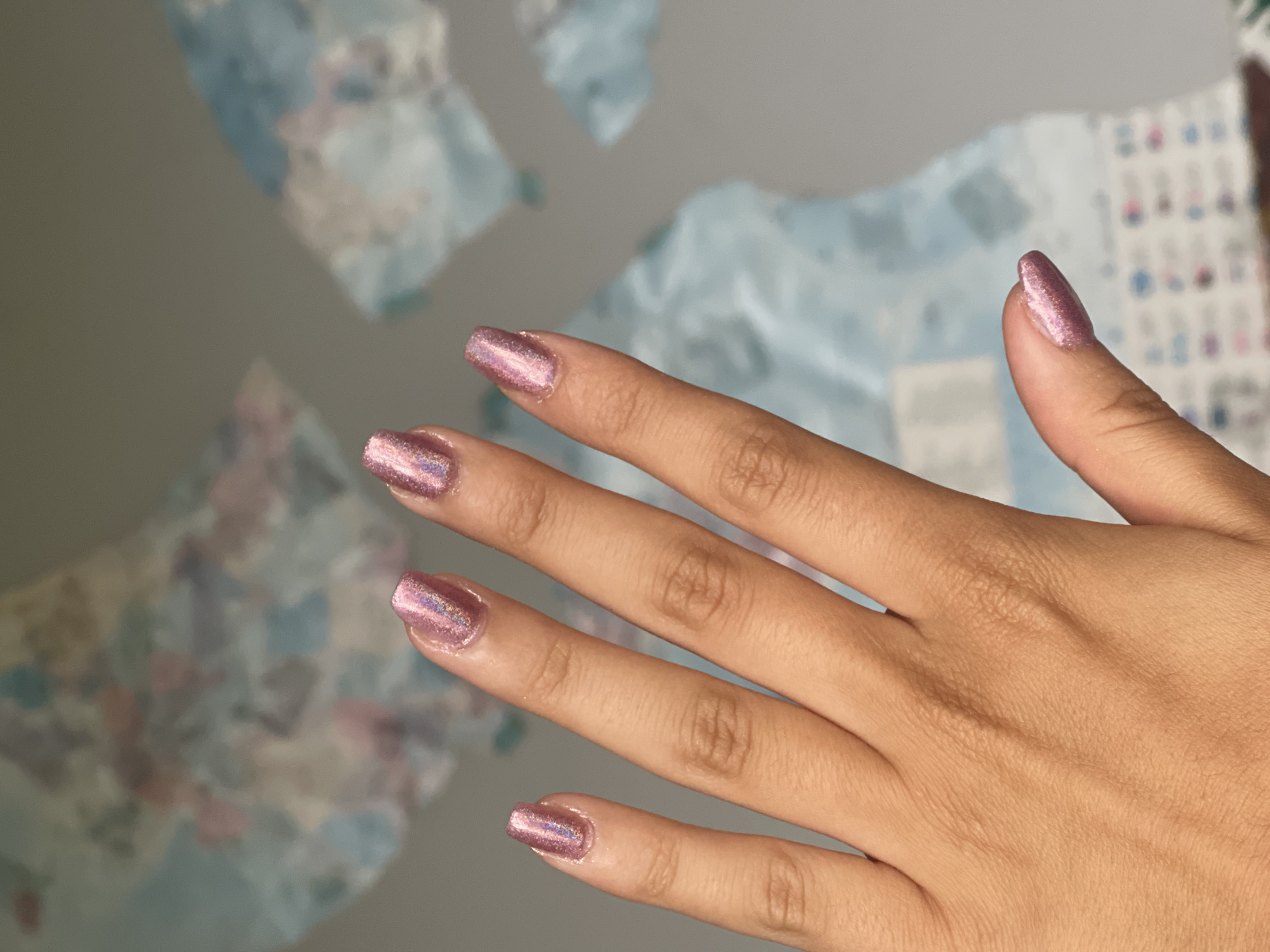 A right human hand with manicured fingernails is positioned against a mottled gray background, with abstract shapes of blue, pink, and white. The fingernails are coated in a rose-gold colored nail polish with a holographic shimmer. The hand is in focus, and the background is slightly blurred. No other people are visible in the image. There is no evidence to suggest that the image was taken in Maidstone, United Kingdom.