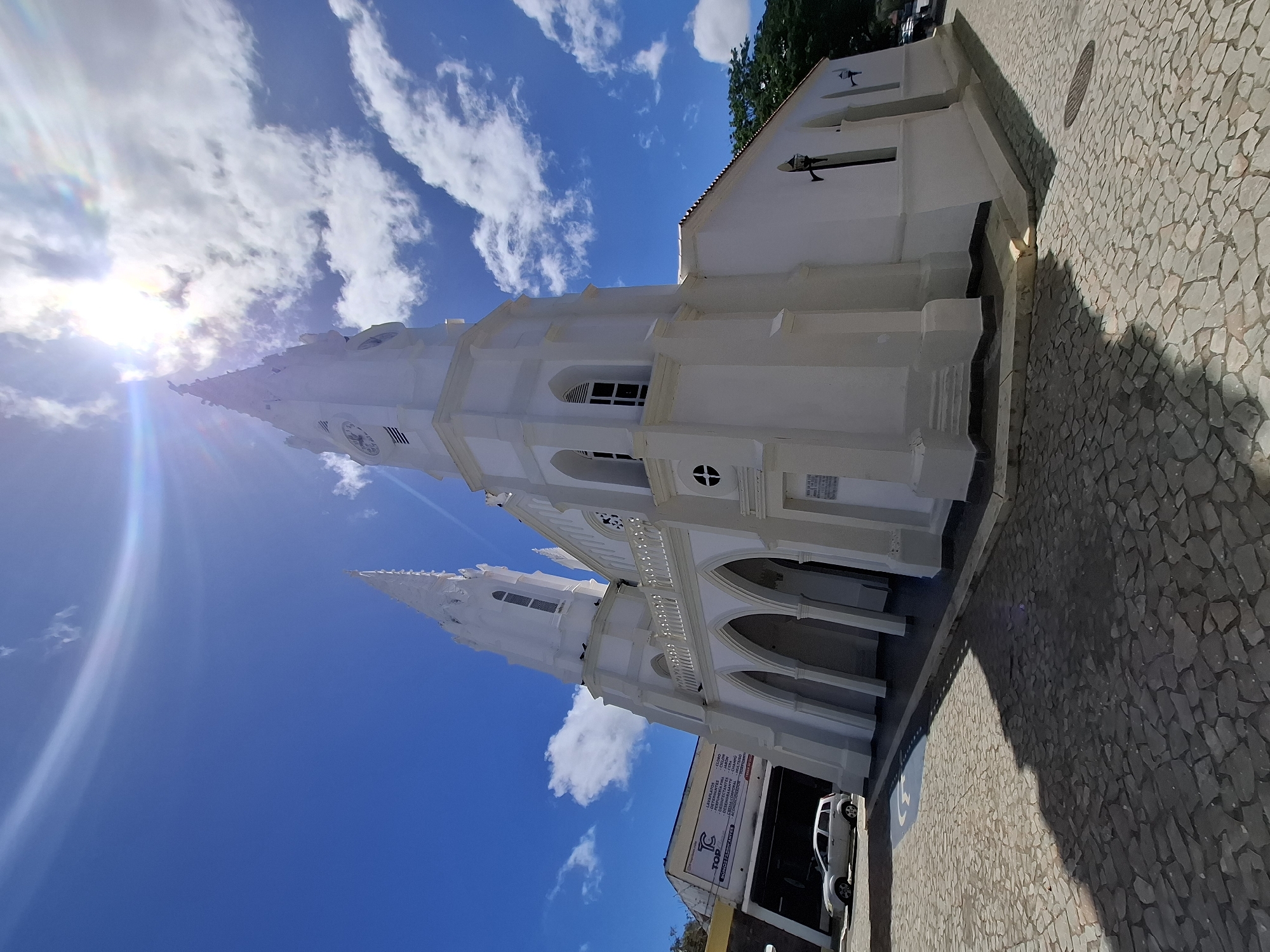 This media file presents a low-angle, rotated view of a striking white church under a vibrant blue sky with scattered white clouds, set in Juan Griego, Venezuela. The dominant subject is a grand, multi-spired church, distinguished by its pristine white facade, tall spires (one featuring a visible clock face), arched doorways and windows, and decorative balustrades.
Intense sunlight from the upper left corner creates a prominent lens flare and casts long shadows across the cobblestone pavement in the foreground and to the right, suggesting it is either late morning or early afternoon. The ground is paved with irregular, light-colored cobblestones.
To the right of the church, a lower building is partially visible. A white car is parked in front of this building, and a large sign is affixed to its facade. The visible text on this sign advertises various products and services, including "TOP", "LAVAPLATO", "DETERGENTE", "CLORO", "PERFUME", "DESINFECTANTE", "FINALABOR", "CERA", "DESENGRASANTE", "CHAMPU", "CREMA CORPORAL", and "MULTIUSO". Below these listings, it states, "SOMOS FABRICANTES" (We are manufacturers).
No people are visible, and no particular event or activity is taking place. The focus is on the architecture of the church and its sunlit surroundings. The bright, clear weather emphasizes the white structure against the deep blue sky.