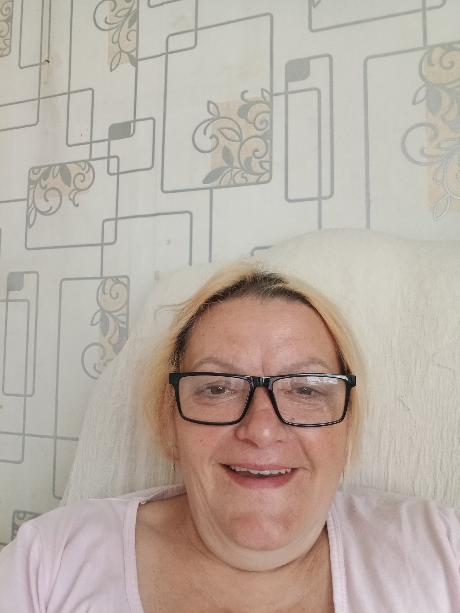 A smiling, middle-aged woman with short, blonde hair is wearing black-framed glasses and a light pink shirt. She appears to be sitting on a white cushion or pillow. The background features a light-colored wallpaper with a geometric pattern interspersed with floral designs. The location is assumed to be in Красный Маяк, Russia.