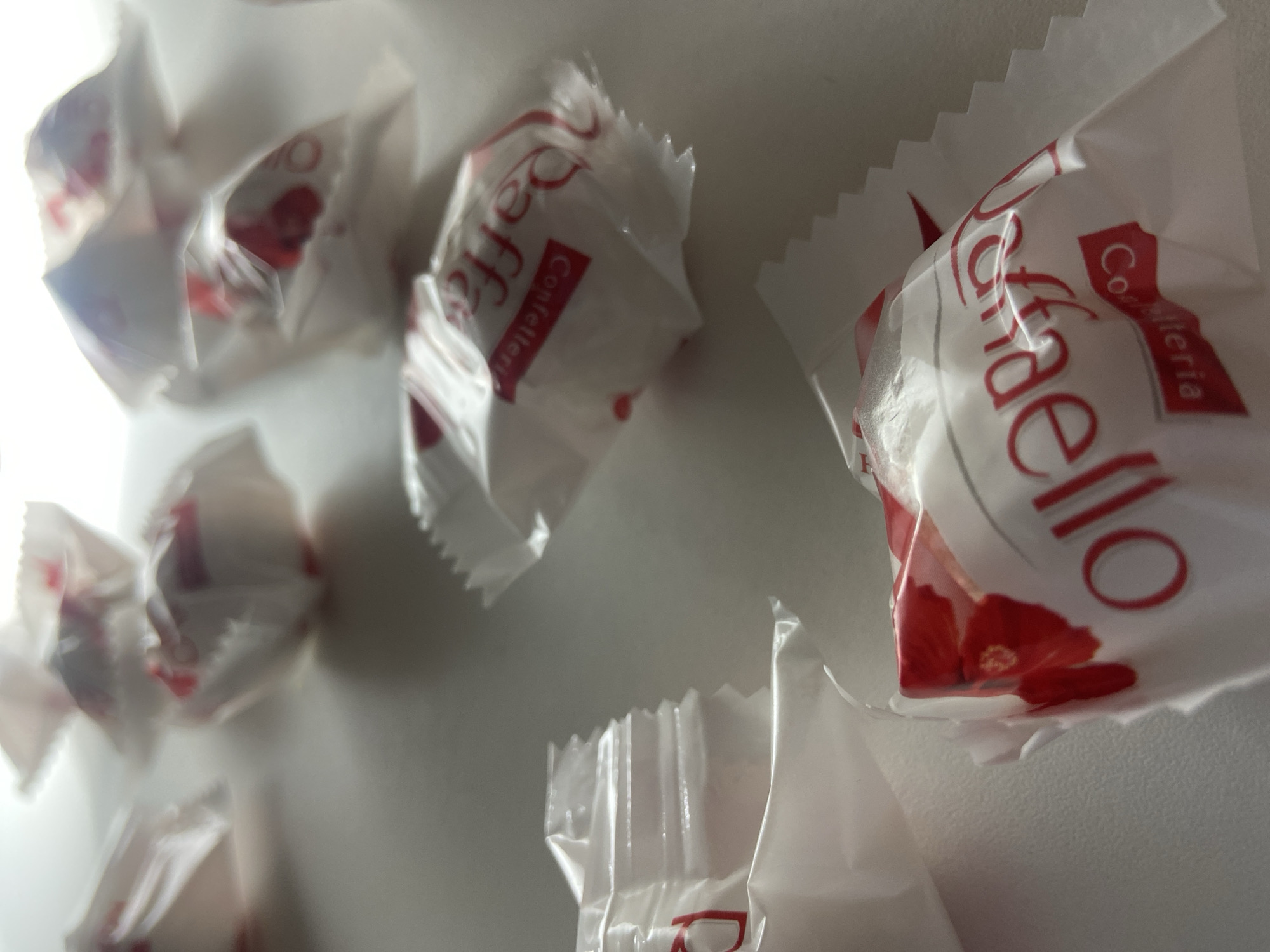 The image displays approximately ten to twelve Raffaello confections scattered on a light-colored, flat surface. Each confection is individually wrapped in white paper with crimped edges, featuring red "Raffaello" branding, "Confetteria" text, and red floral designs. Some wrappers exhibit minor crumpling. The foreground items are in sharp focus, while those in the background are progressively blurred, indicating a shallow depth of field. Bright, diffused light illuminates the scene from an elevated source, casting soft shadows that extend towards the lower right. No individuals, specific actions, or interactive elements are visible. The immediate environment is a simple, uniform surface. The image provides no visual data to identify a specific location within the city of Budva, Montenegro.