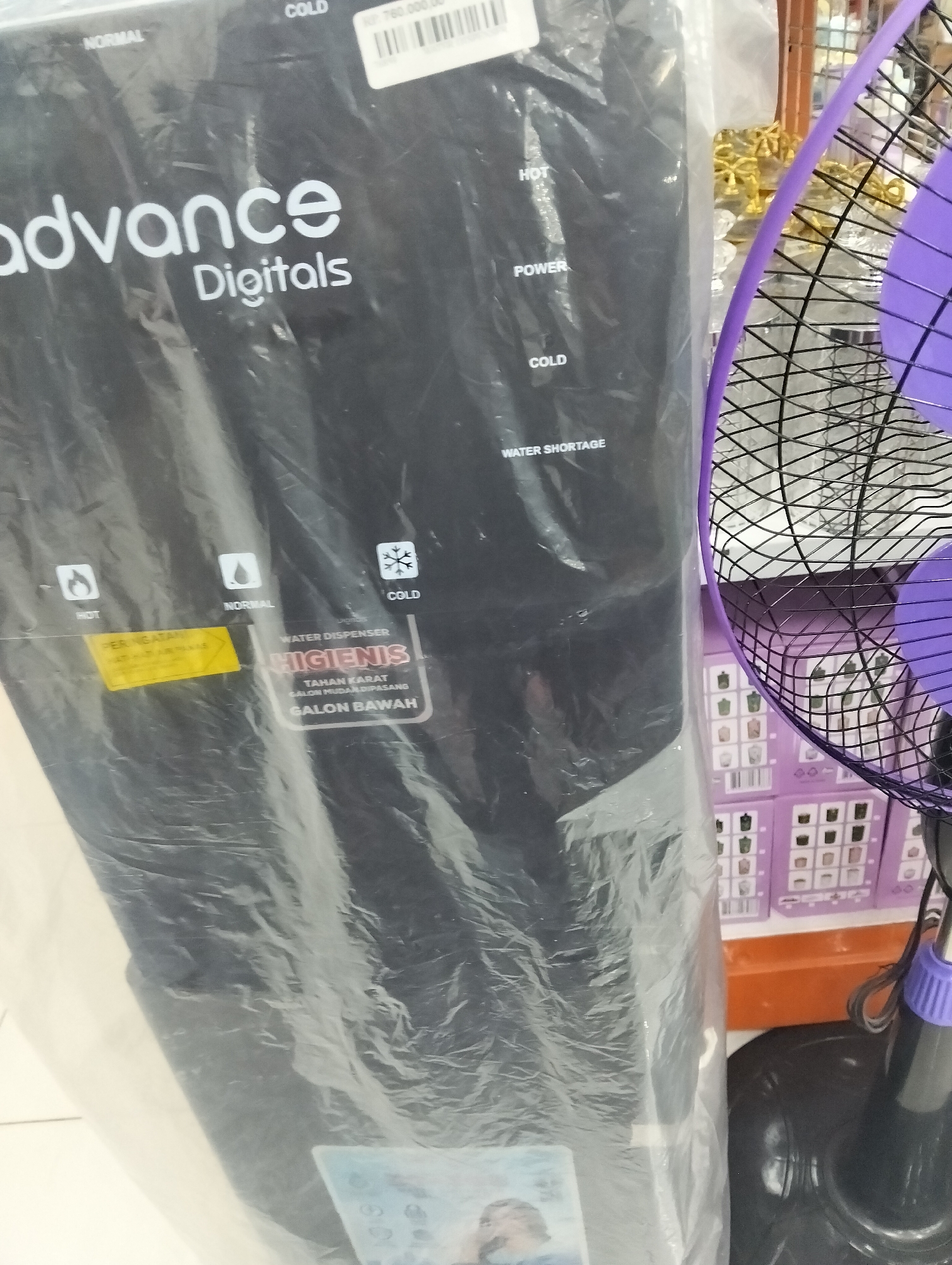 The image shows a close-up of a black water dispenser, branded "advance Digitals", wrapped in clear plastic. The dispenser has indicators for "NORMAL", "COLD", "HOT", "POWER", and "WATER SHORTAGE". There is also a yellow sticker on the dispenser. To the right of the dispenser, a portion of a black and purple electric fan is visible, with its blades and cage clearly discernible. In the background, there are shelves displaying several boxes, some of which appear to contain similar water dispensers or related products, in various shades of purple. An orange shelf is visible below these boxes. The overall scene suggests a retail or storage environment, potentially a shop or warehouse. The lighting is adequate, and the focus is primarily on the water dispenser and the fan. No individuals are visible in the image. The presence of a product with "advance Digitals" branding might suggest it is a retail store in Muncar, Indonesia, where such electronics are sold.
