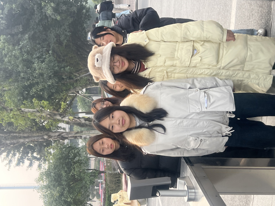 An outdoor scene in Jiangjin, China, depicts a group of predominantly young East Asian individuals, likely forming a queue or standing together. In the immediate foreground, a young woman with dark hair wears a light grey jacket with a prominent light brown fur collar and dark trousers, looking directly towards the observer with a neutral expression. To her right, another young woman with dark hair, wearing round eyeglasses and a light brown fuzzy bucket hat adorned with a bear face, smiles faintly while looking downwards. She is clad in a long, light yellow puffer coat over a striped garment. To the left and slightly behind the first woman, a third young woman with dark hair, wearing a dark puffer jacket, stands with a neutral expression.
Further behind these individuals, several other persons are partially visible. On the far right, a young woman in a dark puffer jacket is visible, and in the distance, a line of people can be observed. To the immediate left of the central foreground figure, a metallic access control gate or turnstile is present. The background features various green trees and bushes, suggesting a landscaped area. Red benches are visible in the mid-ground on the left. A tall building, possibly a skyscraper, is faintly visible in the far background on the left through an apparent atmospheric haze. The ground is paved, and the overall lighting suggests an overcast or hazy condition.