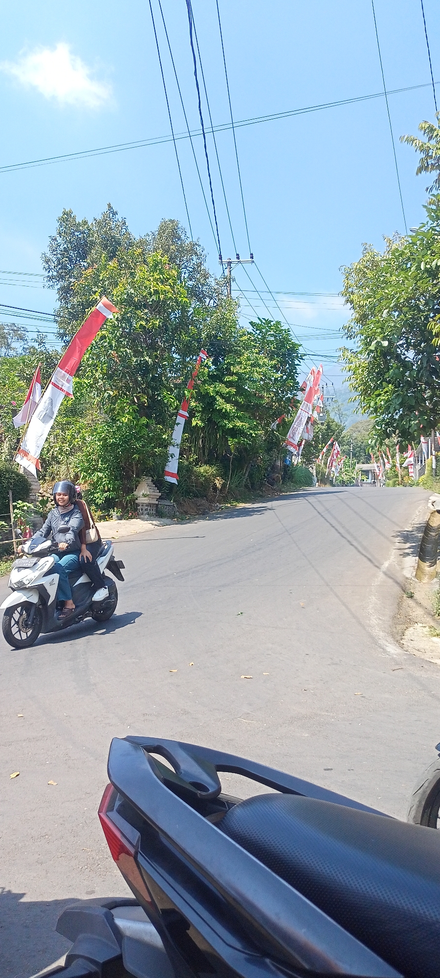 Two individuals are riding a white scooter on a paved road in Selotapak, Indonesia, on a sunny day. The person in the front, wearing a grey long-sleeved shirt, blue jeans, and a black helmet, is operating the scooter. A second person, with dark hair and wearing a brown bag on their back, is a passenger. Red and white flags, likely Indonesian flags, are displayed along the roadside, decorating trees and poles, suggesting a festive occasion or national celebration. The road is lined with lush green trees and vegetation. Overhead, power lines crisscross the clear blue sky. In the foreground, the back portion of another dark-colored scooter is visible, partially obscuring the scene. The road appears to be a rural or semi-rural area, with a gentle incline upwards into the distance.