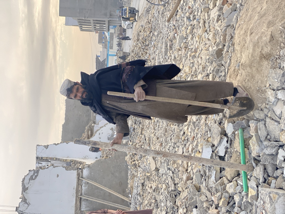 This image captures a street scene in Kandahar, Afghanistan, characterized by extensive rubble and partially demolished structures, indicative of a demolition site or an area affected by destruction.
In the foreground, a middle-aged male individual with a dark beard and mustache stands prominently on a small mound of dirt amidst a vast expanse of concrete and brick debris. He wears a brown shalwar kameez, a dark embroidered shawl draped over his shoulders, and a white, textured cap (possibly a pakol). He holds a long, dirt-covered wooden pole vertically in his left hand, and a shovel, blade-down, in his right hand. His gaze is directed towards the viewer. To his immediate left, a portion of another person, wearing a brown garment, is partially visible. Two green pipes lie horizontally on the rubble near the foreground.
In the mid-ground, behind the main subject, are the remains of a multi-story building, its white-painted facade significantly damaged, exposing internal brickwork, rebar, and supporting beams. Another smaller structure with dark openings is visible behind this ruined building. Further back, on the right side of the frame, a paved road with active traffic is visible. Vehicles include several yellow and white auto-rickshaws, one or more trucks, and a pedestrian on a bicycle. A modern, multi-story building, possibly under construction or recently completed, stands along the road on the right.
The background features a hazy, light-colored sky with scattered clouds, and distant hills or mountains are visible on the horizon. The overall lighting suggests late afternoon or early evening.