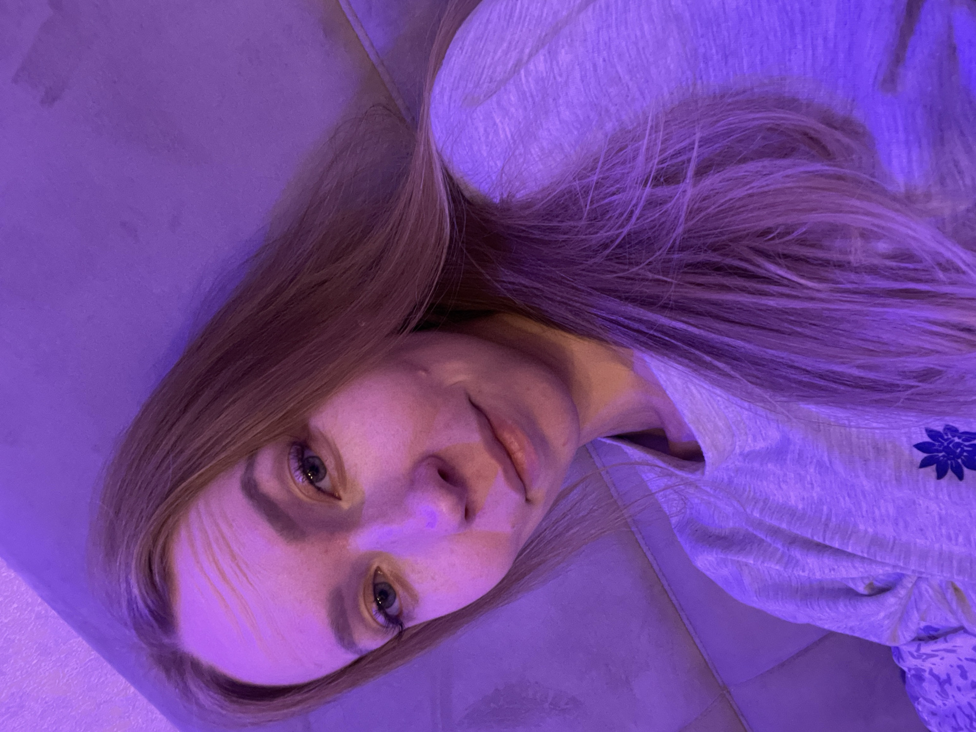 A young Caucasian woman with blonde hair is lying down, facing the camera at an angle. She is wearing a light-colored t-shirt. The lighting is heavily tinted purple. The location appears to be indoors, possibly in a private residence. There is no verifiable information to definitively place the image in Perm, Russia.