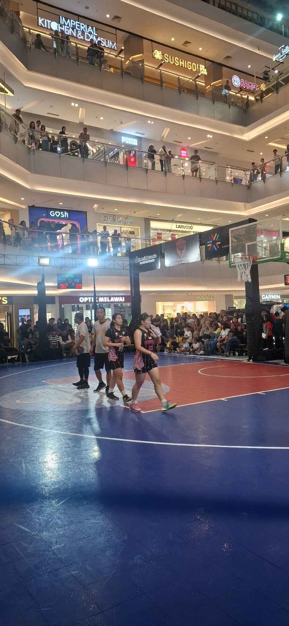 The image depicts an indoor basketball court within a multi-level shopping mall in Grogol, Indonesia. The court is marked with blue and red boundaries. Several people are visible on the court, including three individuals in athletic attire standing near the center line. One person is holding a basketball. In the background, a large crowd of spectators is gathered, observing the activity. Above the court, various retail store signs are illuminated, with "GOSH" and "OPTIC MELAWAI" being legible. The mall's architecture features multiple levels with railings and escalators. A basketball hoop and backboard are positioned on the right side of the frame. The overall scene suggests a sporting event or exhibition taking place within a commercial setting.