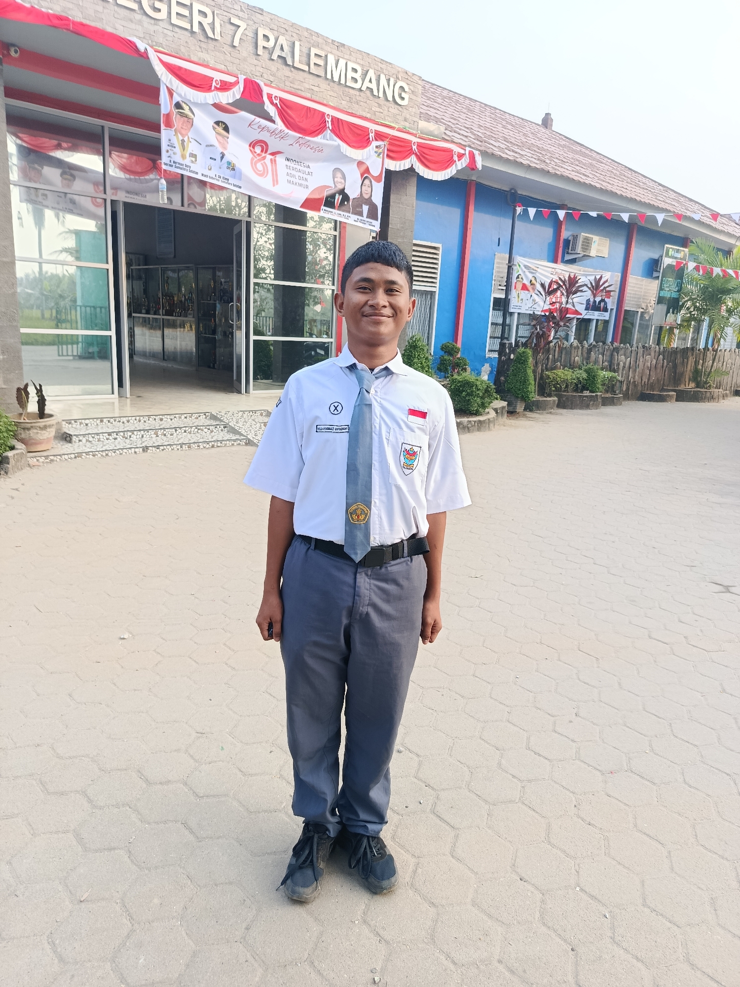 A young man in a white shirt, gray pants, and a tie stands in front of a blue building with the words "NEGERI 7 PALEMBANG" written above the entrance. The building is decorated with red and white banners and flags. The man is smiling and looking at the camera. He appears to be a student at the school. The ground is paved with hexagonal tiles. The background shows trees and a fence. The scene is outdoors and appears to be during the daytime.