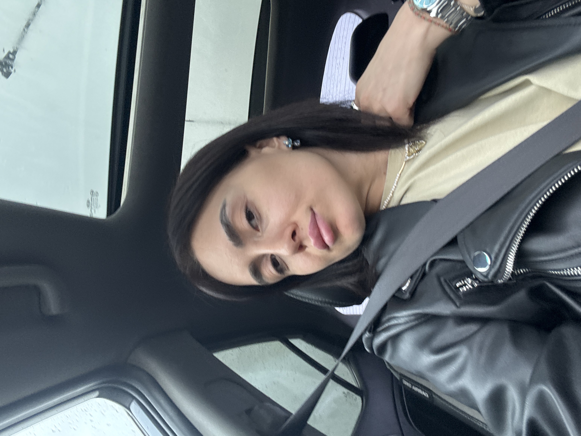 A single adult female is depicted in a close-up, chest-up shot, seated in the front passenger seat of a vehicle. She has dark hair and is wearing a black leather jacket over a light beige t-shirt. A black seatbelt is diagonally across her chest and right shoulder. Accessories include a pearl necklace, a small pendant, a silver-toned wristwatch with a turquoise-colored bezel on her left wrist, a multi-strand bracelet of red and brown beads on the same wrist, and a blue and silver earring on her visible left ear. Her left hand is positioned near her collarbone. The car interior components are dark grey or black. A "SRS AIRBAG" label is visible on the pillar to the viewer's left. A sunroof and a rear side window are visible, both covered in water droplets, suggesting rain and an overcast sky. The individual is looking directly at the camera. The image contains no visual information to determine a specific location within Tbilisi, Georgia.